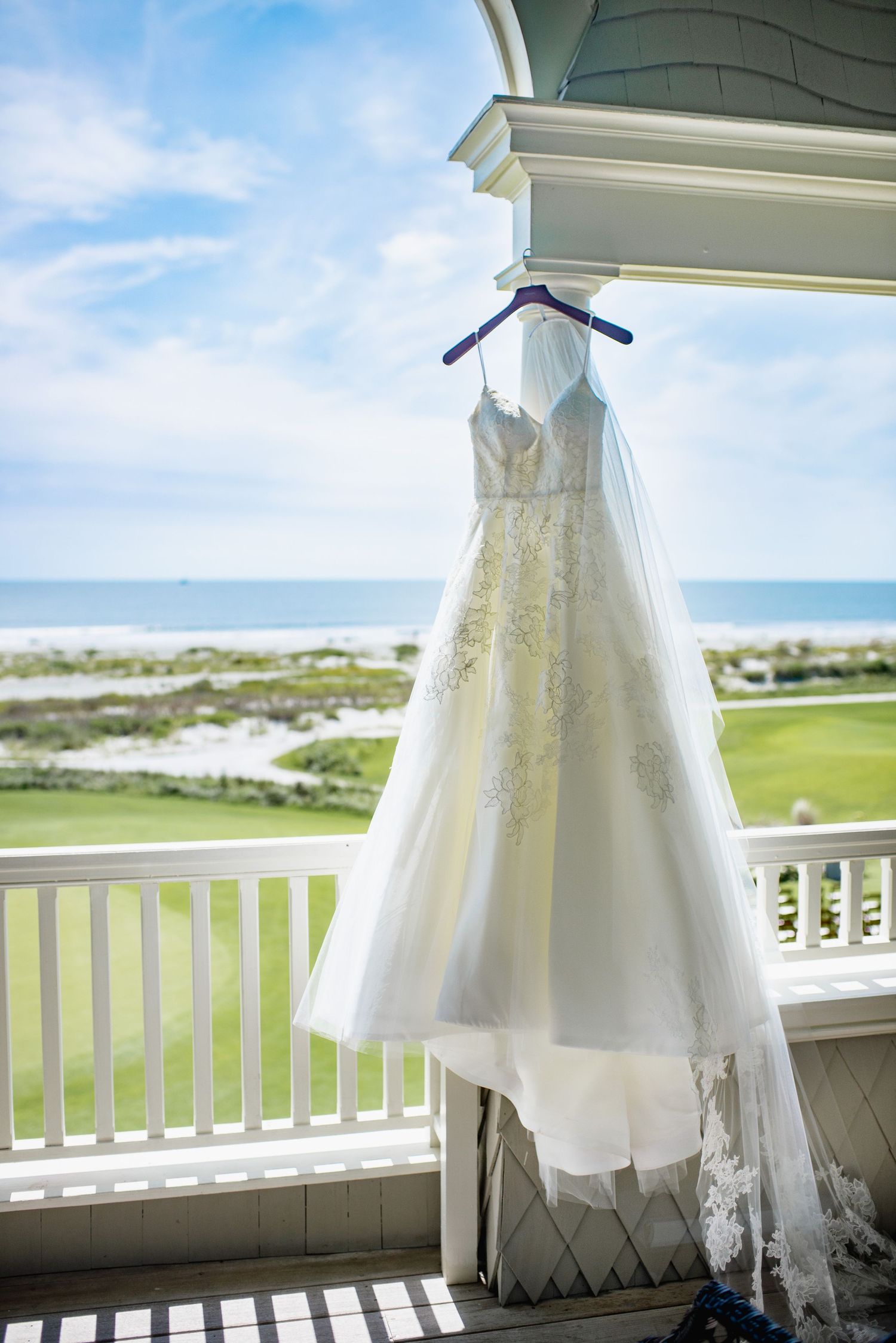 A white lace wedding dress hangs from a balcony railing overlooking a scenic coastal landscape on a sunny day.