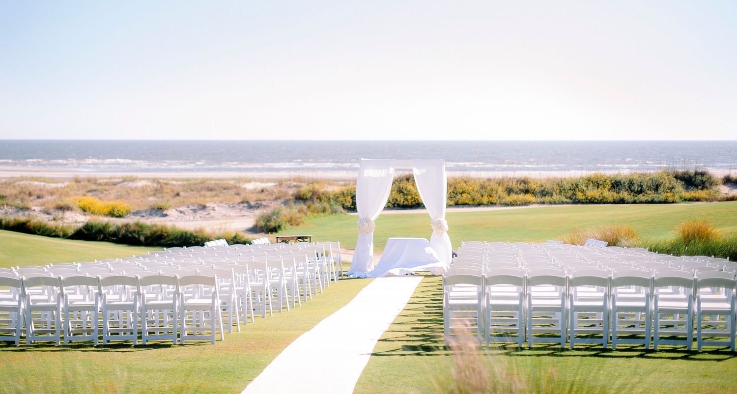 White wedding ceremony setup with chairs and arch overlooking a scenic coastal landscape on a sunny day.