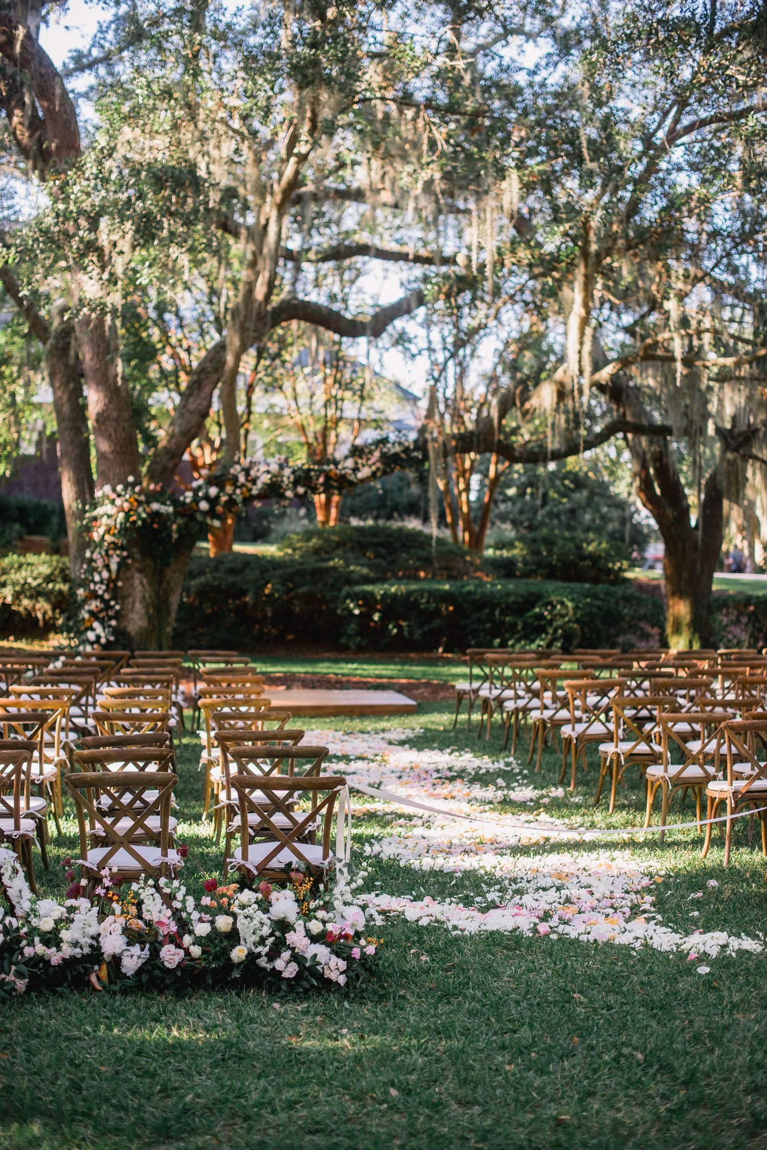 Wooden chairs arranged in rows for an outdoor wedding ceremony with white flower petals scattered along a grass aisle.