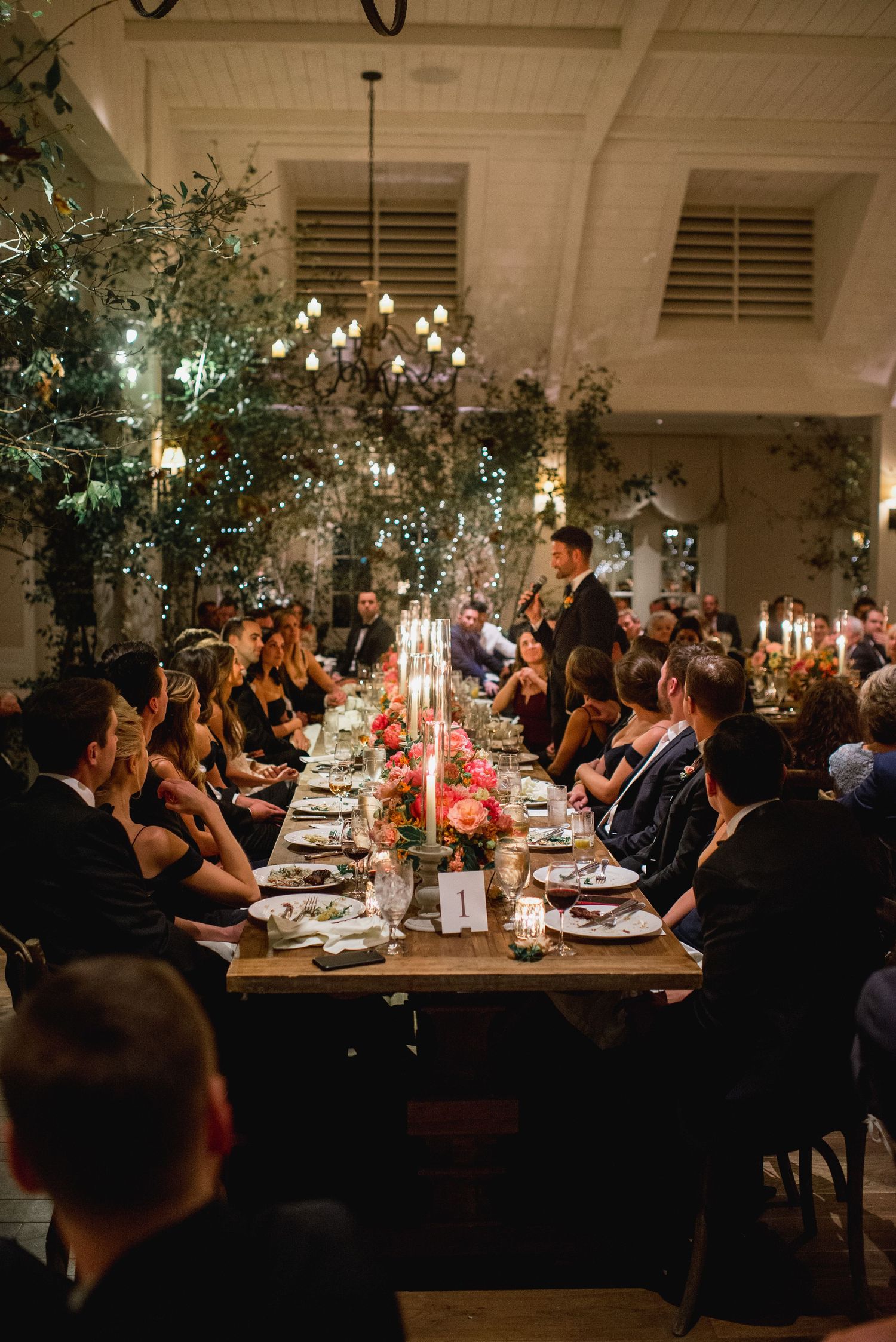 Long dining tables set with candles and greenery create an intimate atmosphere at an indoor wedding reception.