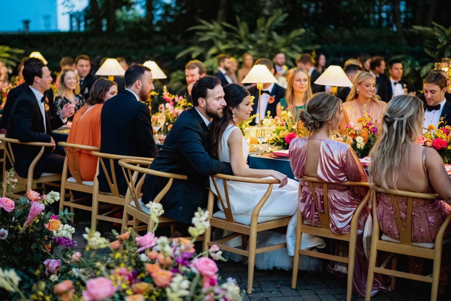 Wedding guests seated at outdoor reception tables with string lights and pink floral arrangements in a garden setting.