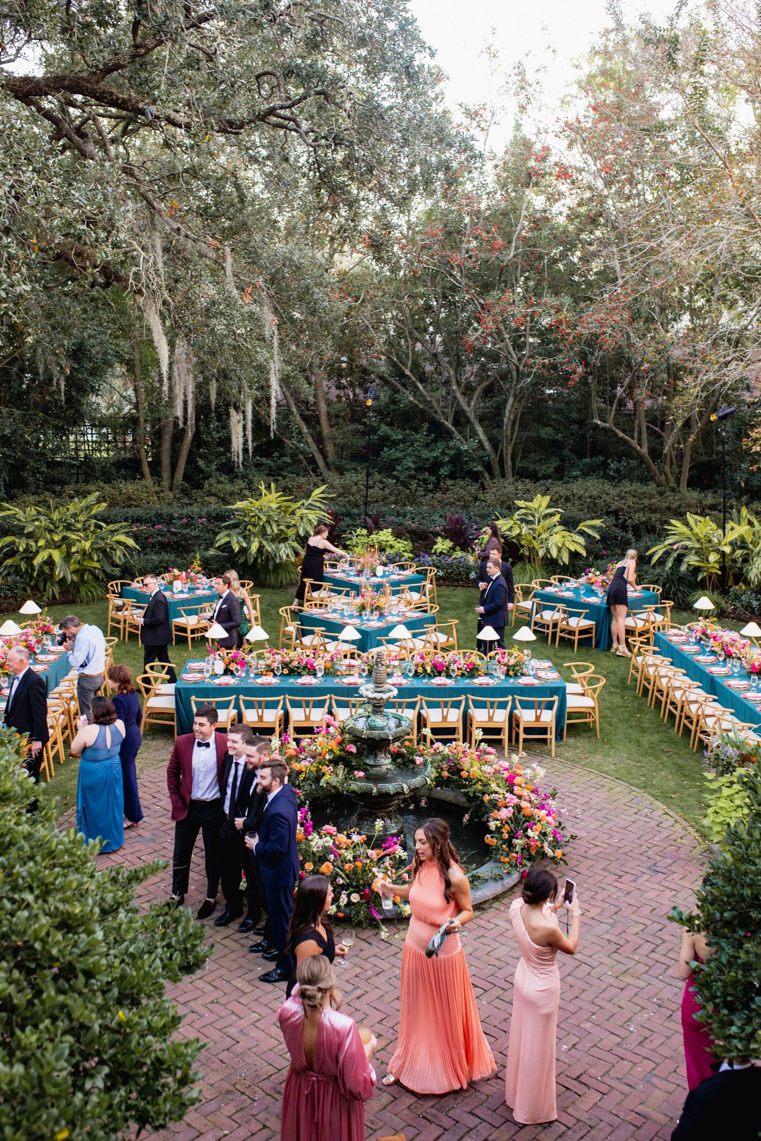 Outdoor garden wedding reception setup with floral centerpieces, long tables, and guests mingling at sunset.