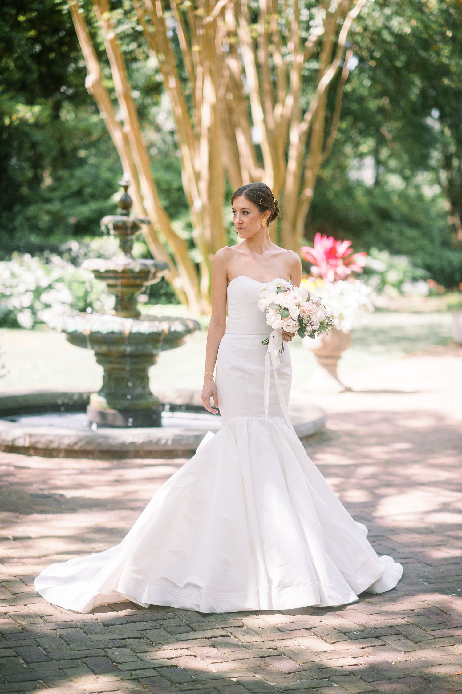 A bride in a classic white ball gown poses gracefully on stone pavers surrounded by lush garden trees.