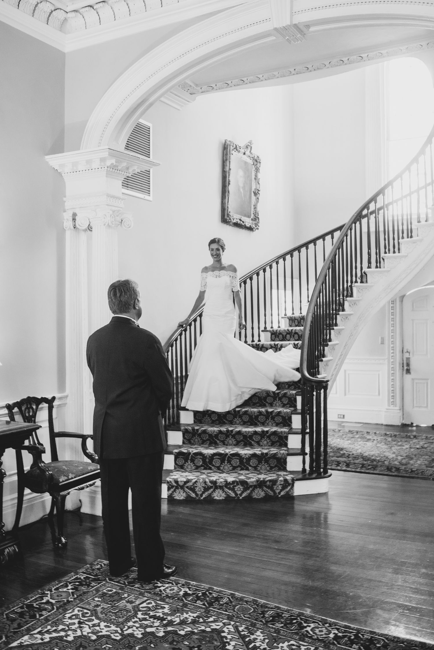 Black and white photo of an elegant staircase in a historic mansion with ornate architectural details and patterned carpet.
