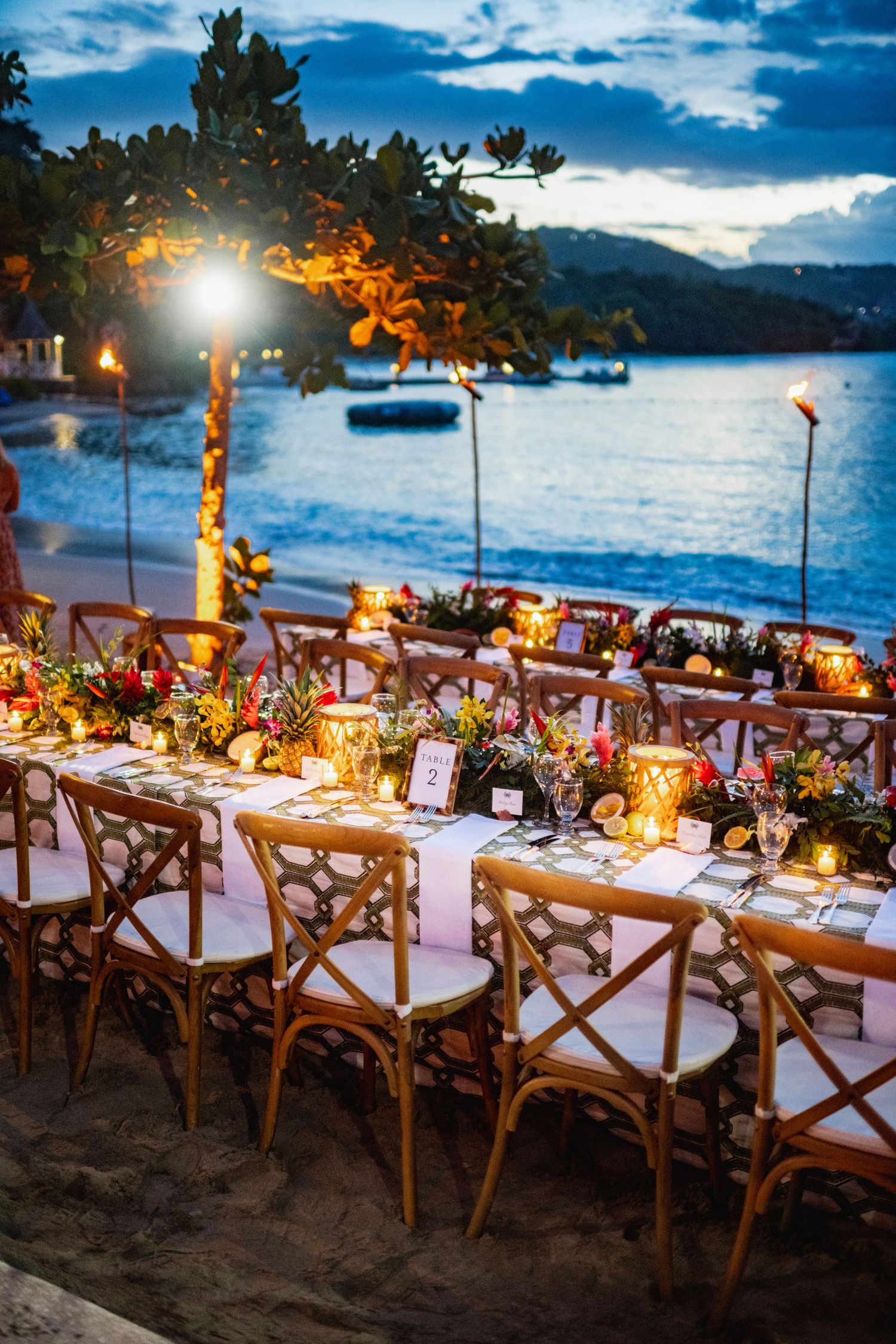 Elegant beach wedding reception tables set with wooden chairs and glowing candles along the shoreline at sunset.