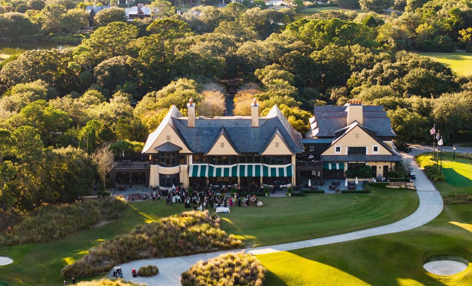 Aerial view of a luxurious golf clubhouse with winding driveway surrounded by lush green trees and manicured grounds.