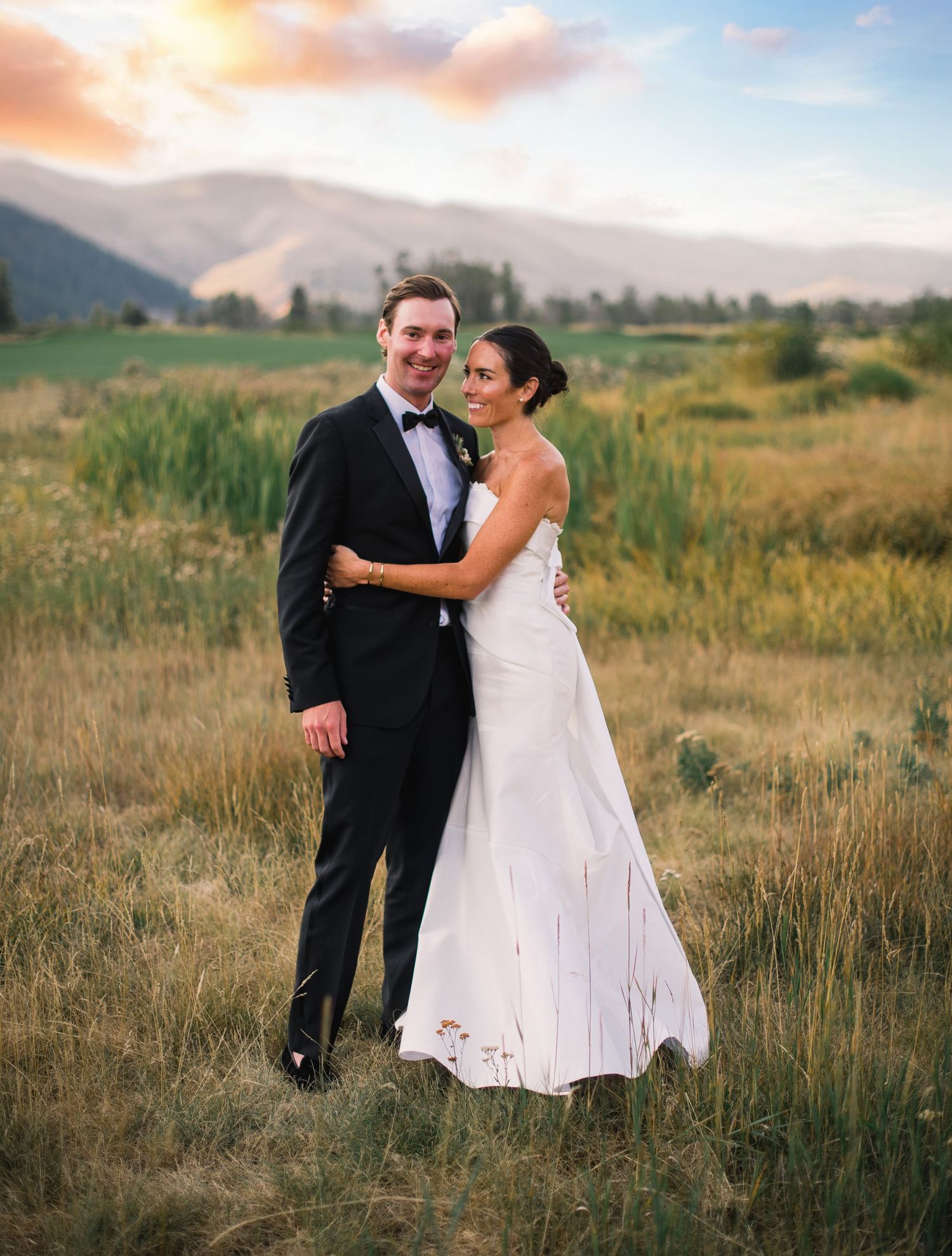 A couple shares a romantic moment in a field at sunset with mountains in the background.
