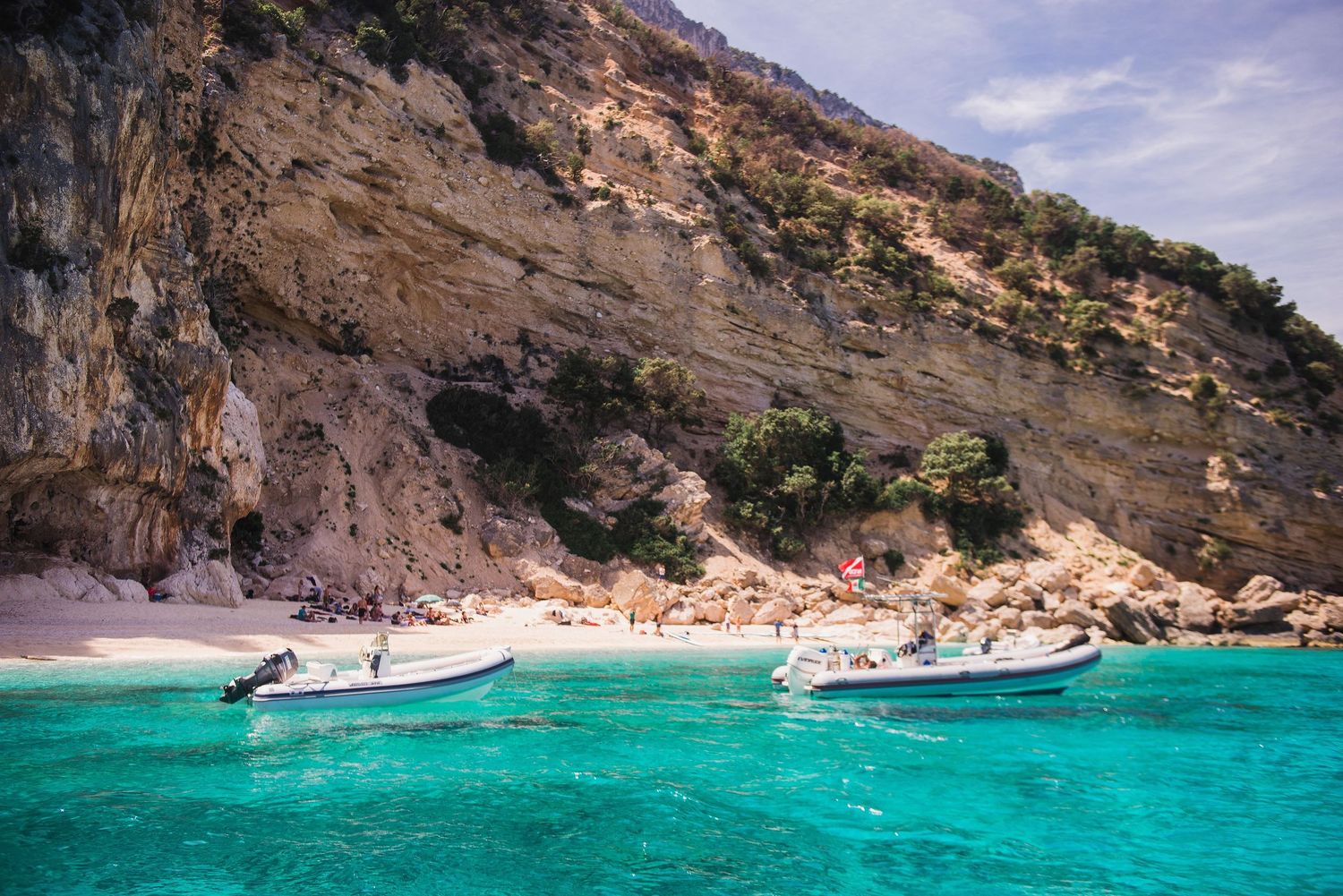 Small boats anchored in crystal clear turquoise waters near a secluded beach surrounded by limestone cliffs.
