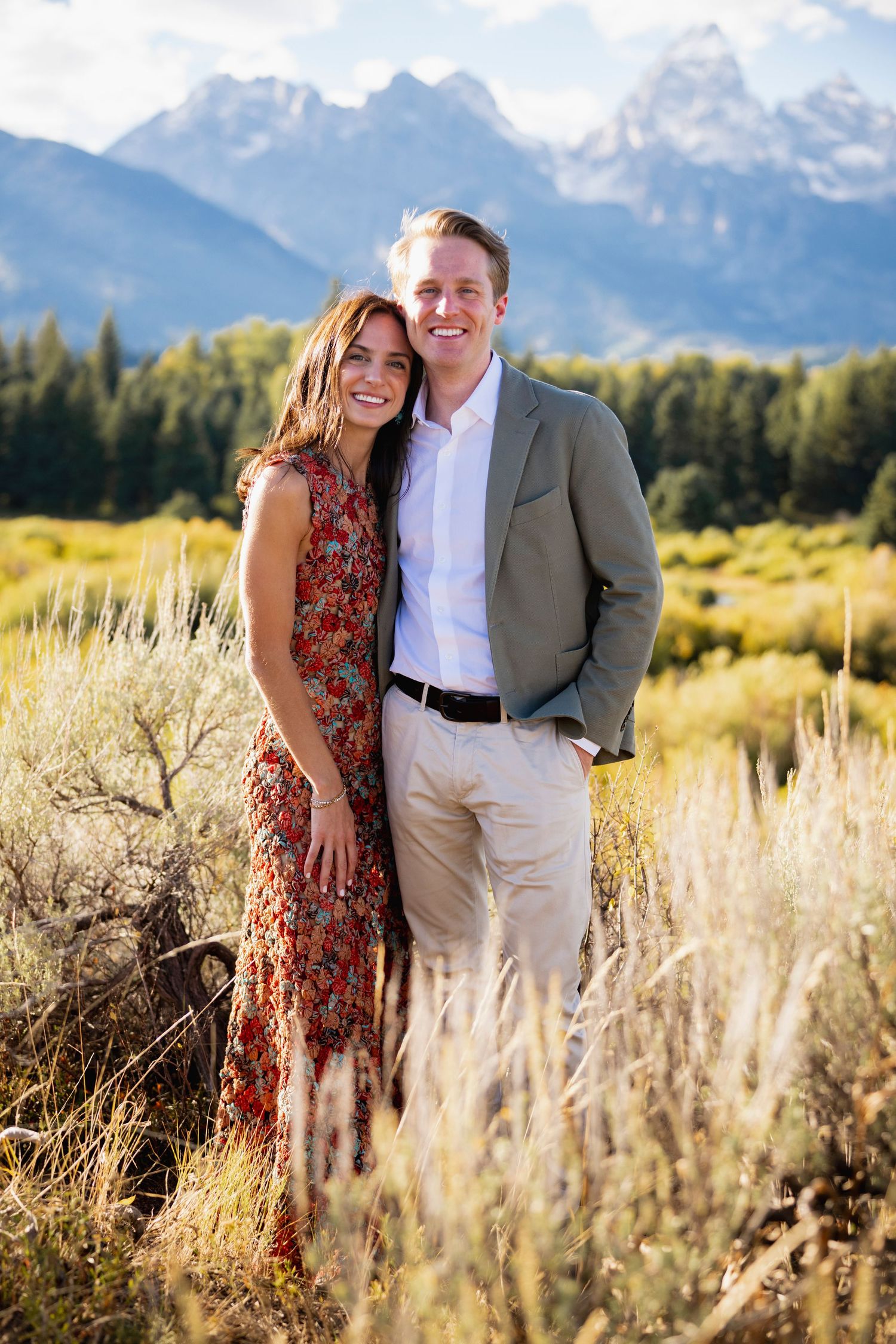 Couple embracing in mountain meadow with snow-capped peaks and golden grass in background.