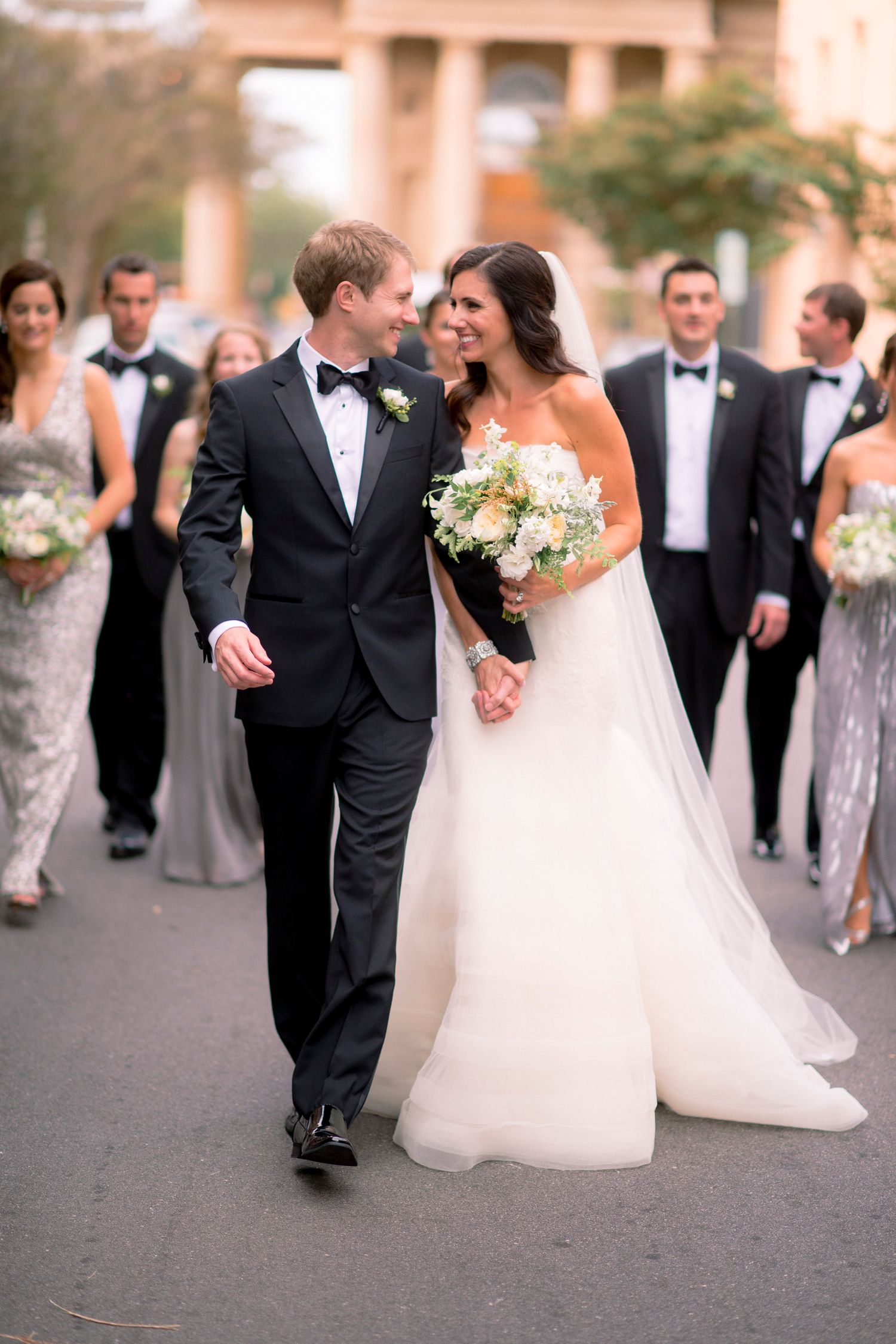 Wedding party walks together on paved path with bride in white gown and groomsmen in black tuxedos.