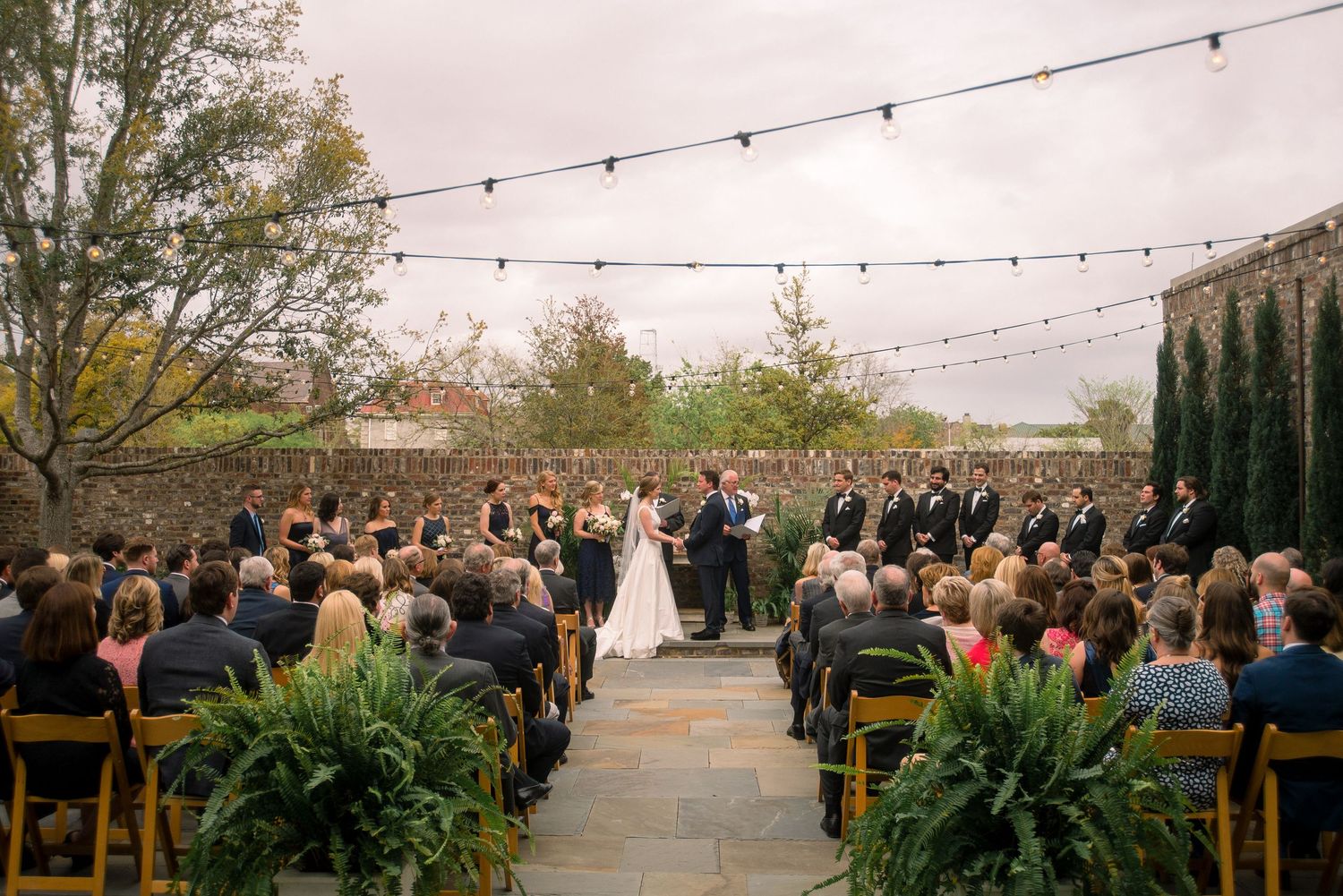 String lights illuminate an outdoor wedding ceremony with guests seated in rows beneath a cloudy desert sky.