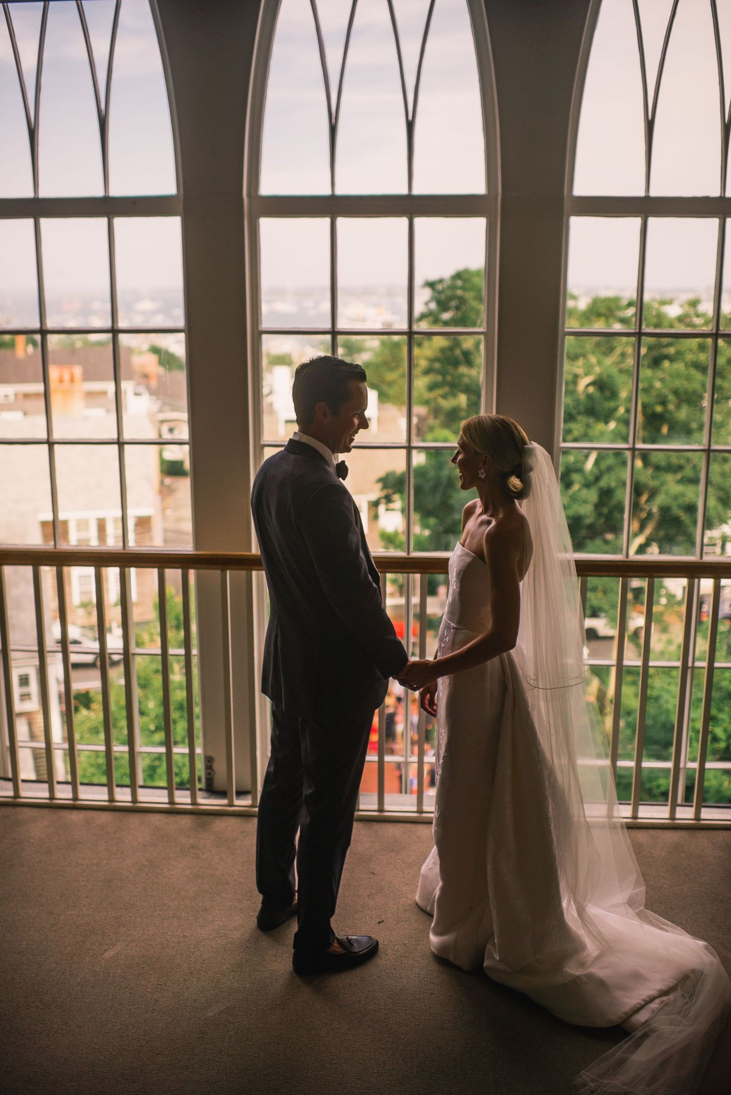Silhouetted couple in wedding attire standing by large windows with city view.