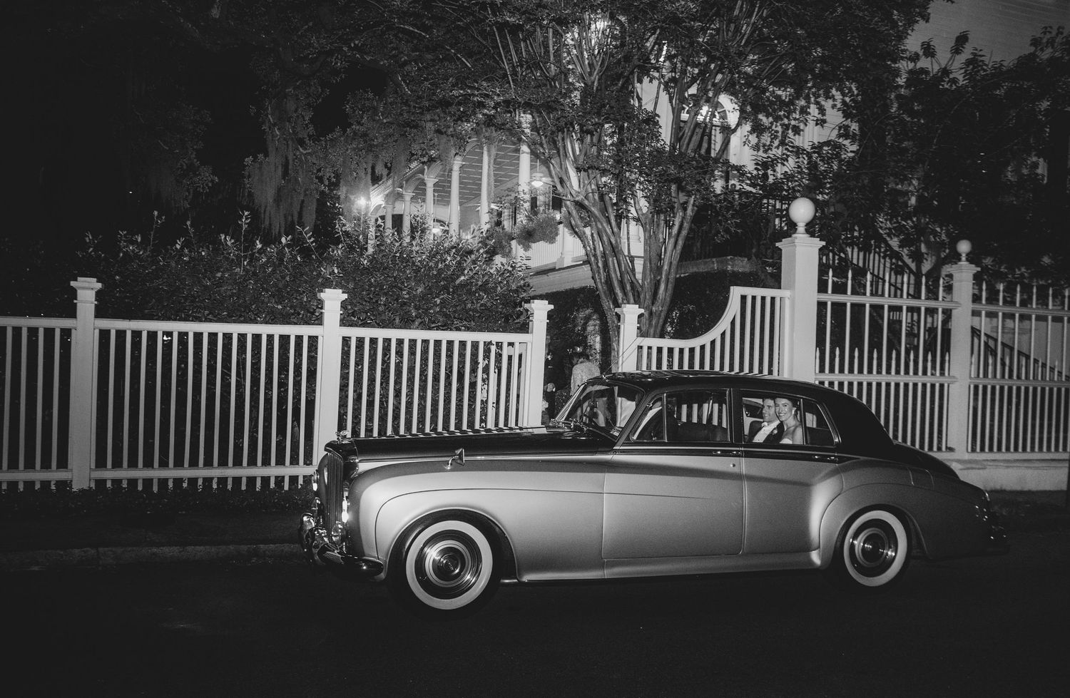 Classic vintage car parked on a tree-lined street with white picket fences in the background.