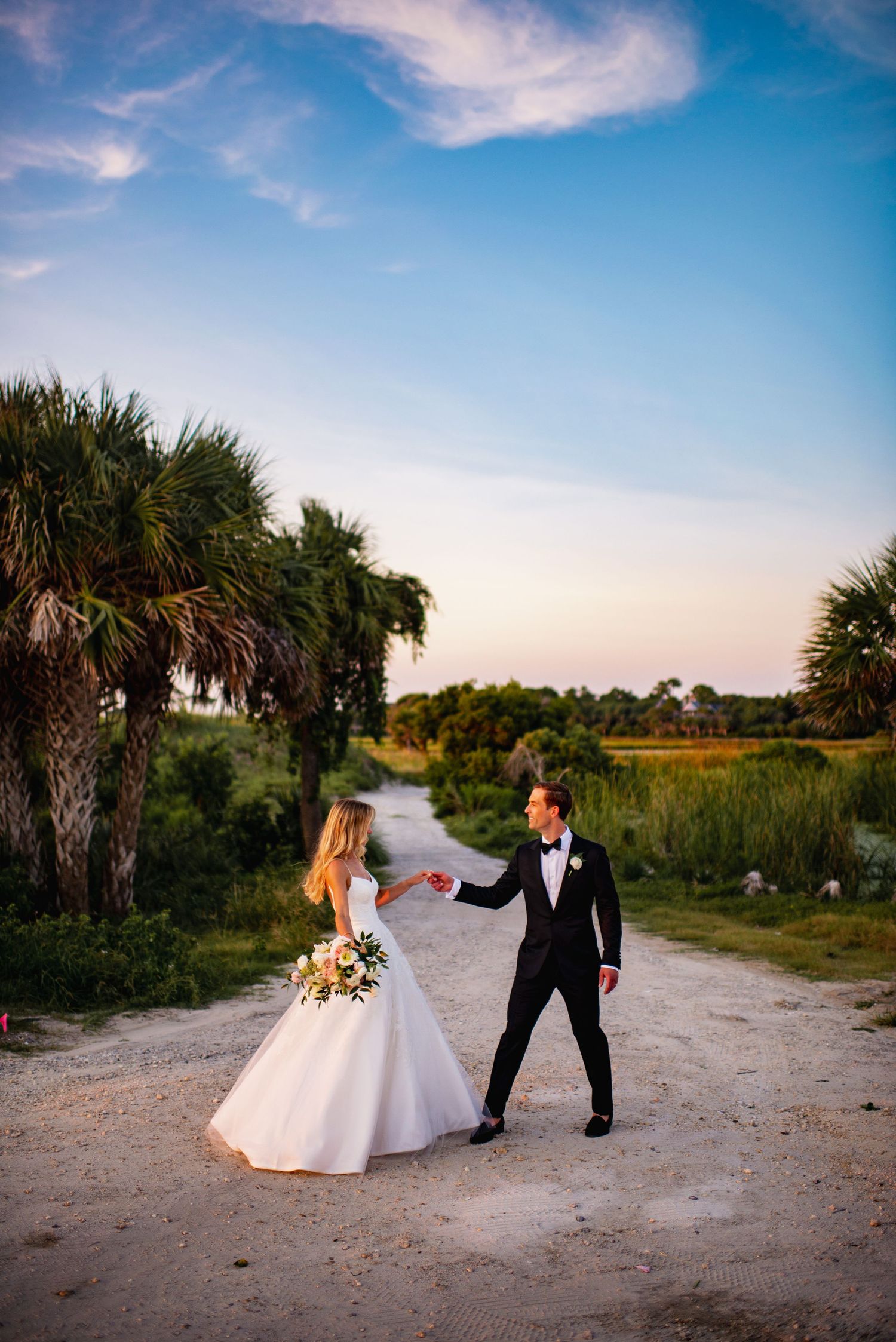 A couple dances together on a sandy beach path surrounded by palm trees and coastal vegetation at sunset.