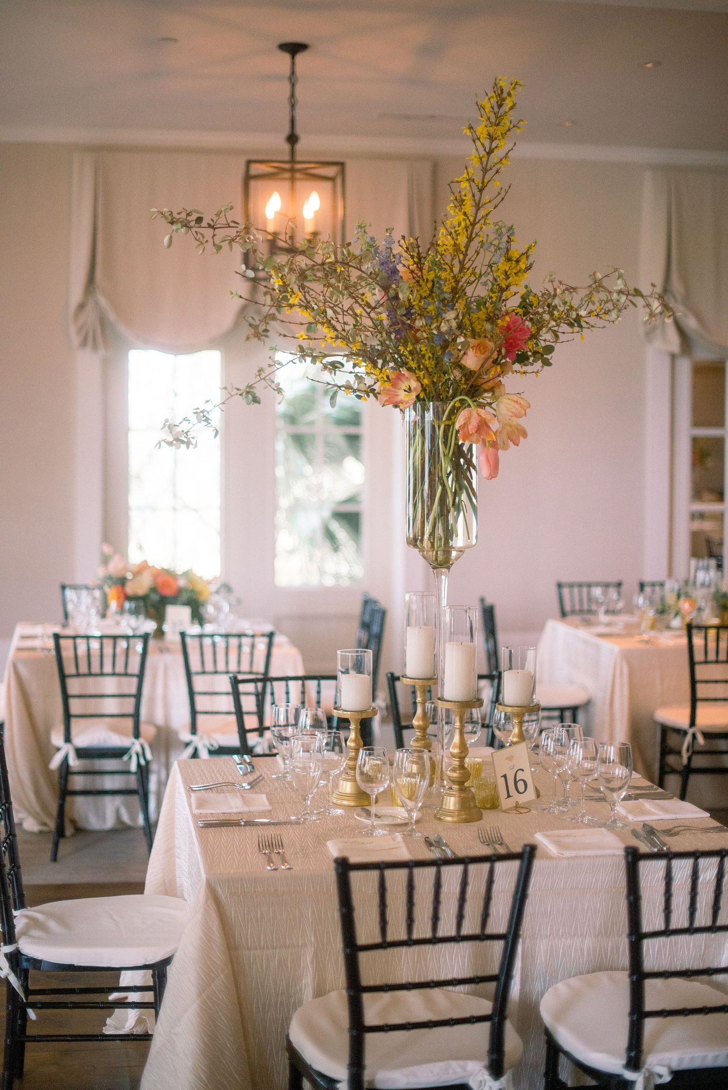 Elegant wedding reception table with tall floral centerpiece, white linens, and black chiavari chairs in bright dining room.