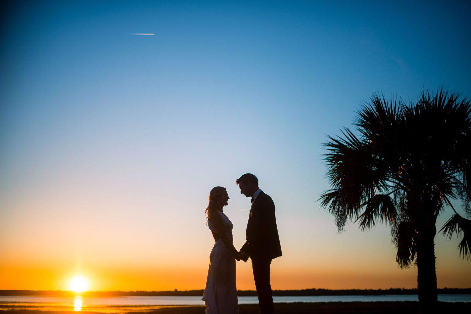 Silhouetted figures walk along a beach at sunset with palm trees and vibrant orange sky in the background.