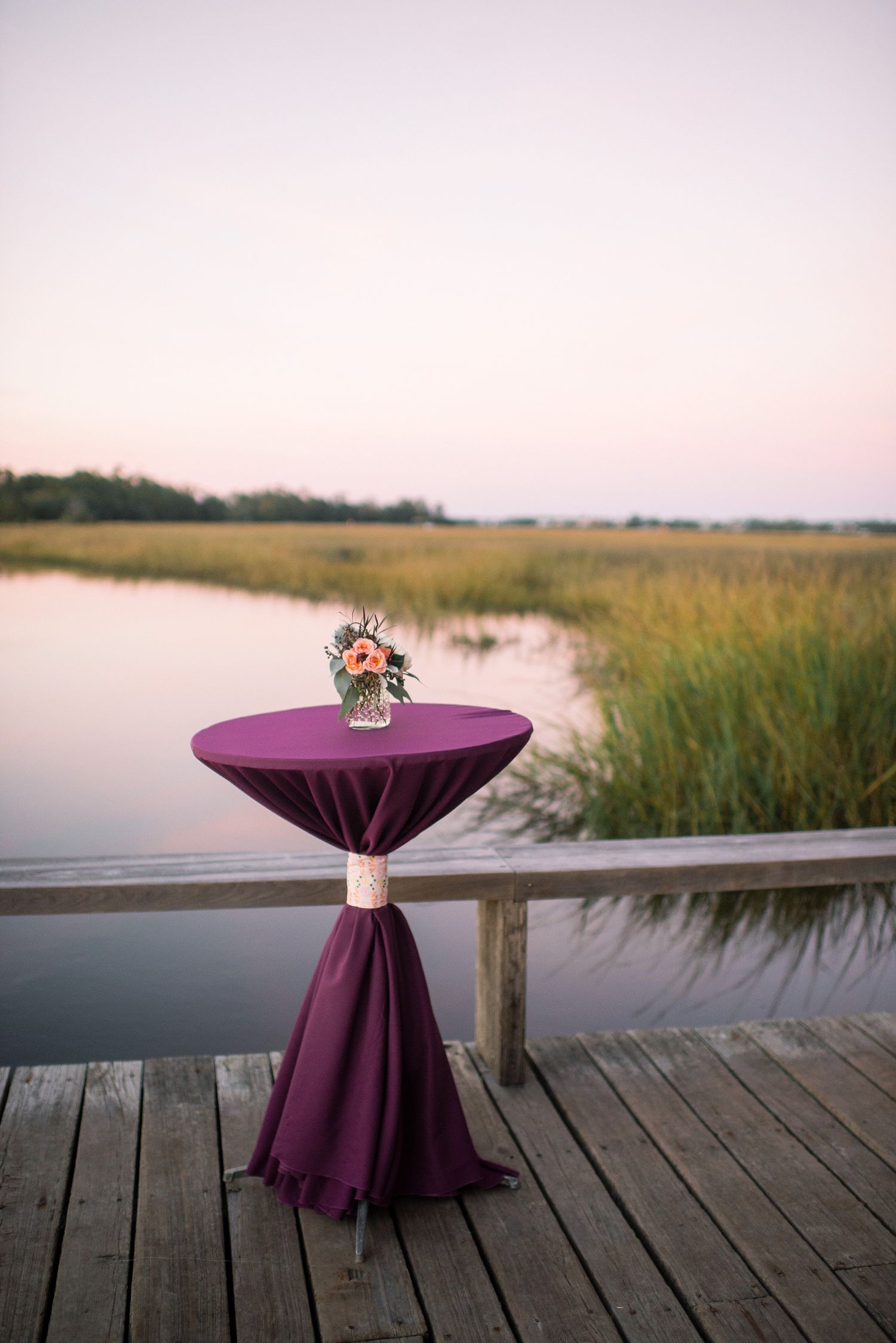 Purple tablecloth-covered cocktail tables sit on a wooden dock overlooking a serene marsh at sunset.