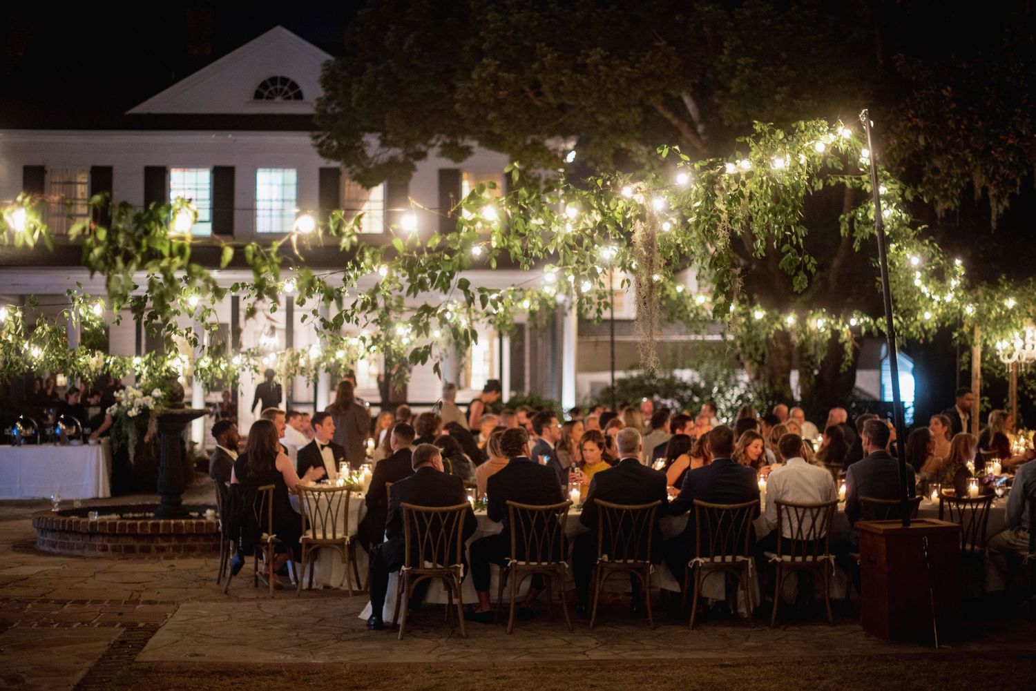 Evening outdoor reception with string lights illuminating long dining tables beneath majestic trees.
