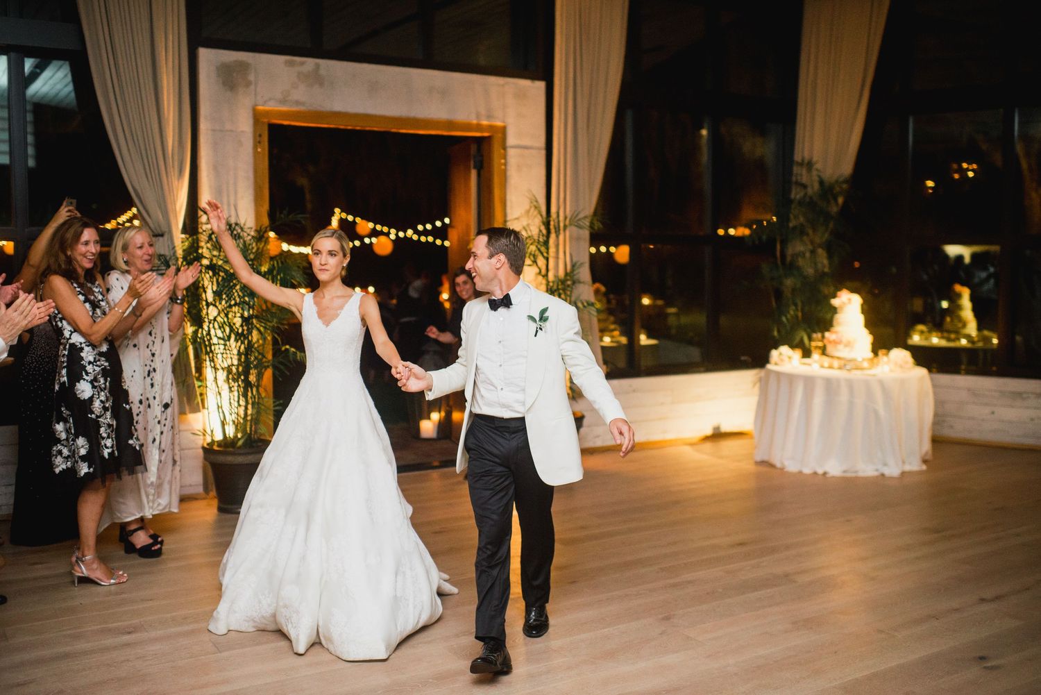 Newlyweds and guests dancing together on a wooden dance floor at an evening wedding reception.