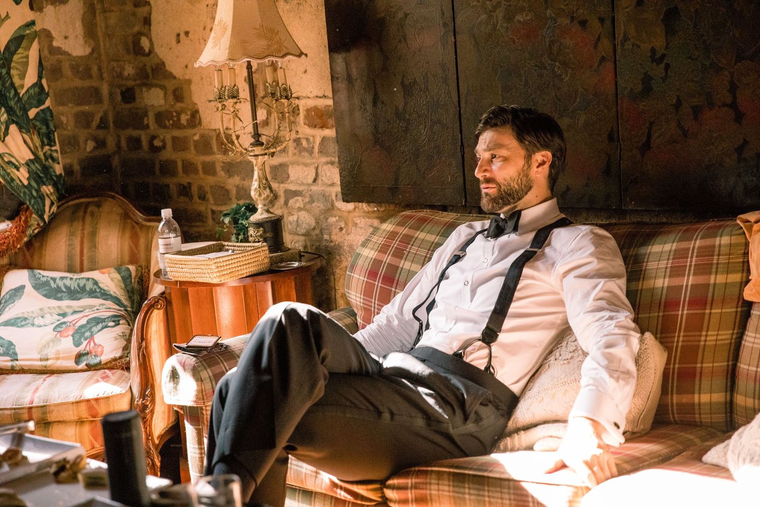 Relaxed man in white dress shirt and suspenders reclines on plaid couch in rustic brick-walled room.