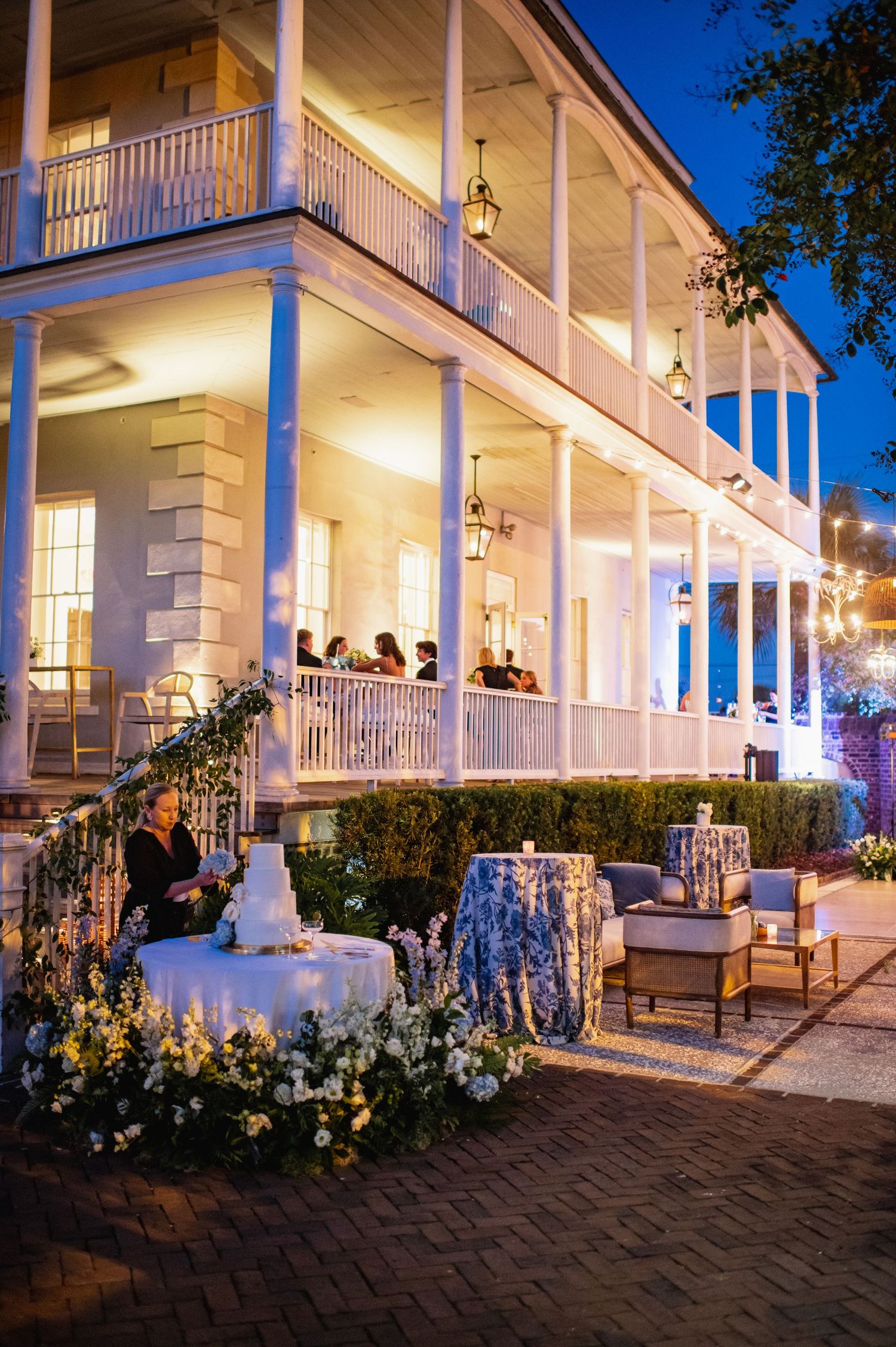 Elegant white colonial-style building with illuminated wraparound porch and brick walkway at dusk.