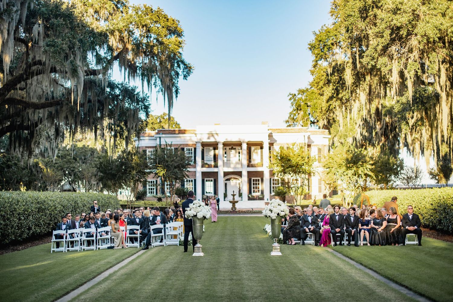 Panoramic view of an elegant outdoor wedding ceremony setup with white chairs on a lush green lawn surrounded by oak trees.