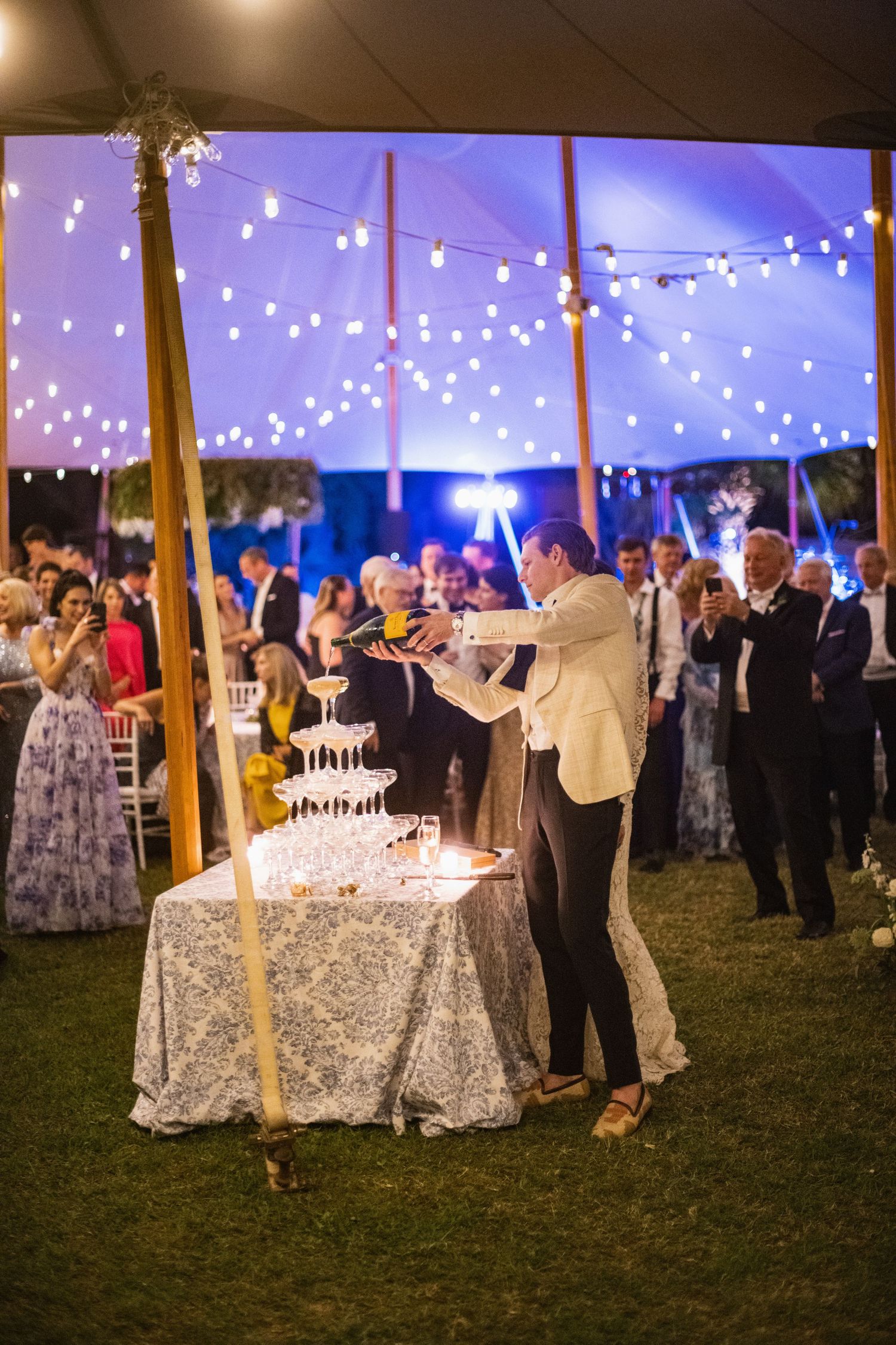 Wedding guests dance and celebrate under a white tent with twinkling lights during an evening reception.