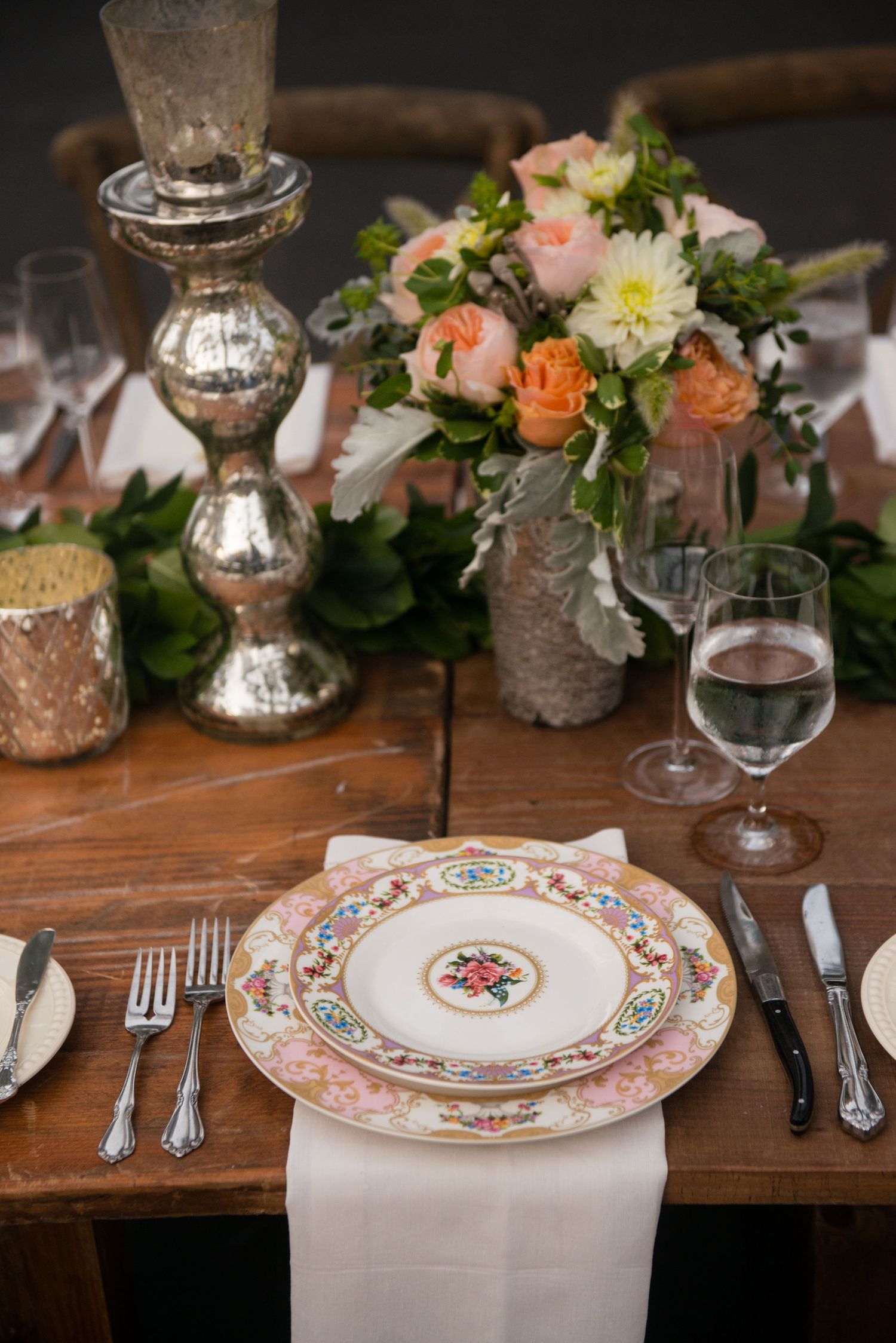 Elegant table setting with vintage floral plates, silverware, and fresh flower centerpieces on rustic wooden tables.