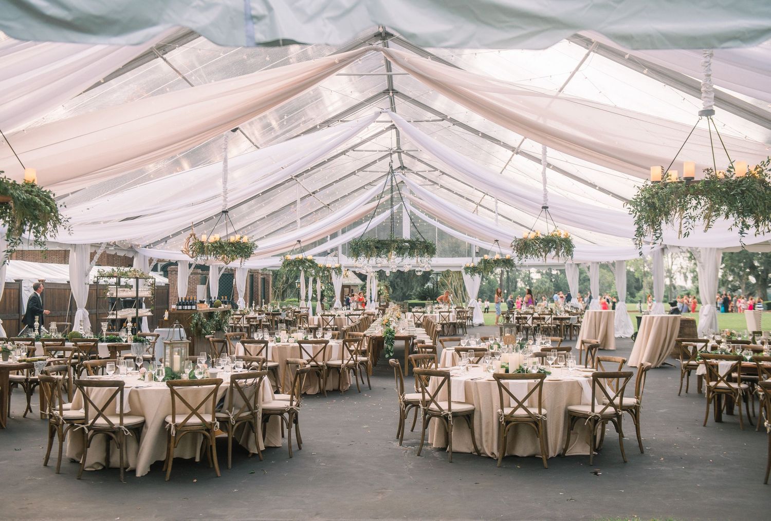 An elegant outdoor wedding reception setup with white tent canopy and cross-back chairs arranged at round tables.