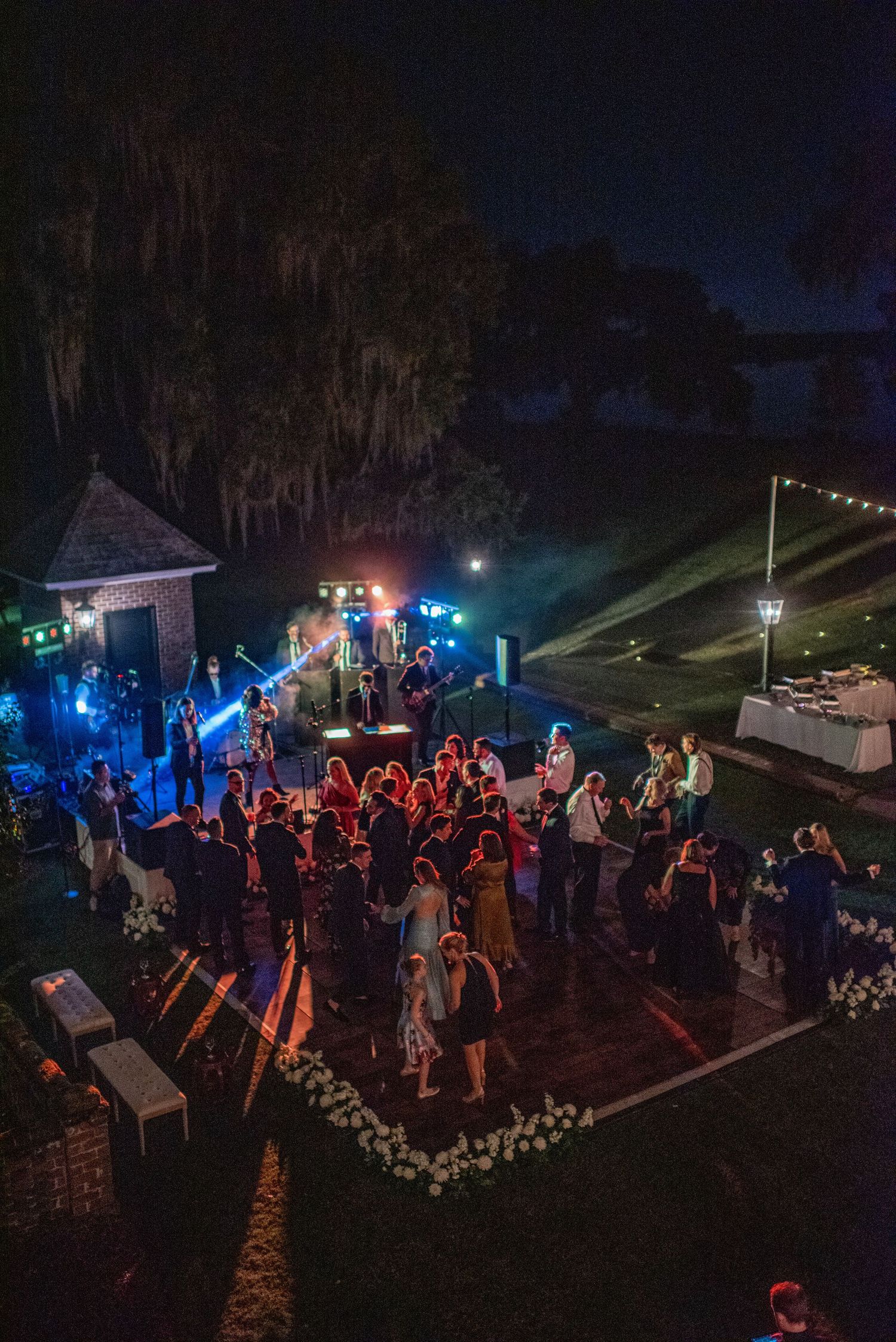 Outdoor nighttime wedding reception dance floor illuminated with blue and red lights as guests dance under trees.