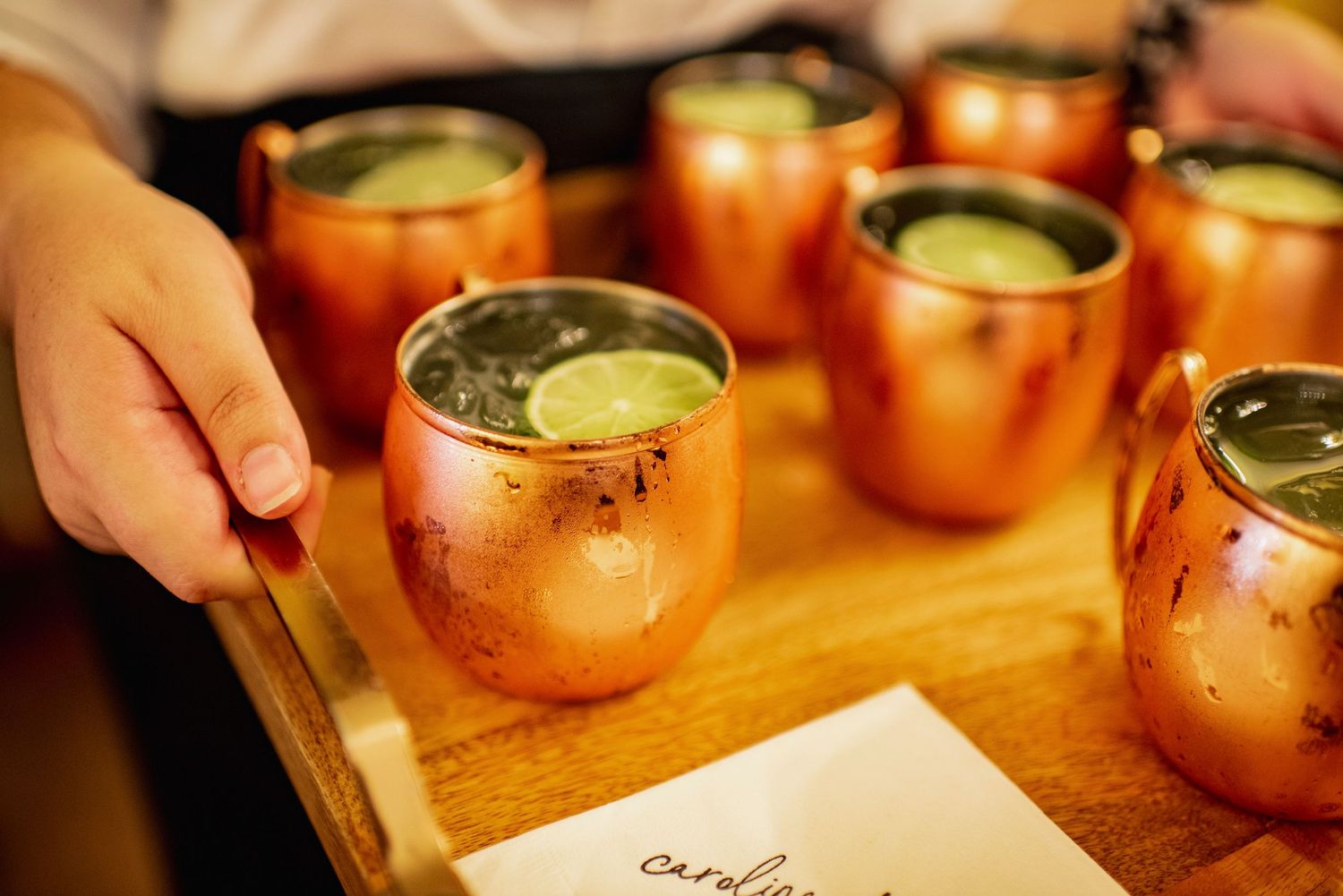 Moscow mule cocktails being poured into copper mugs with lime garnish and ginger beer on a wooden surface.