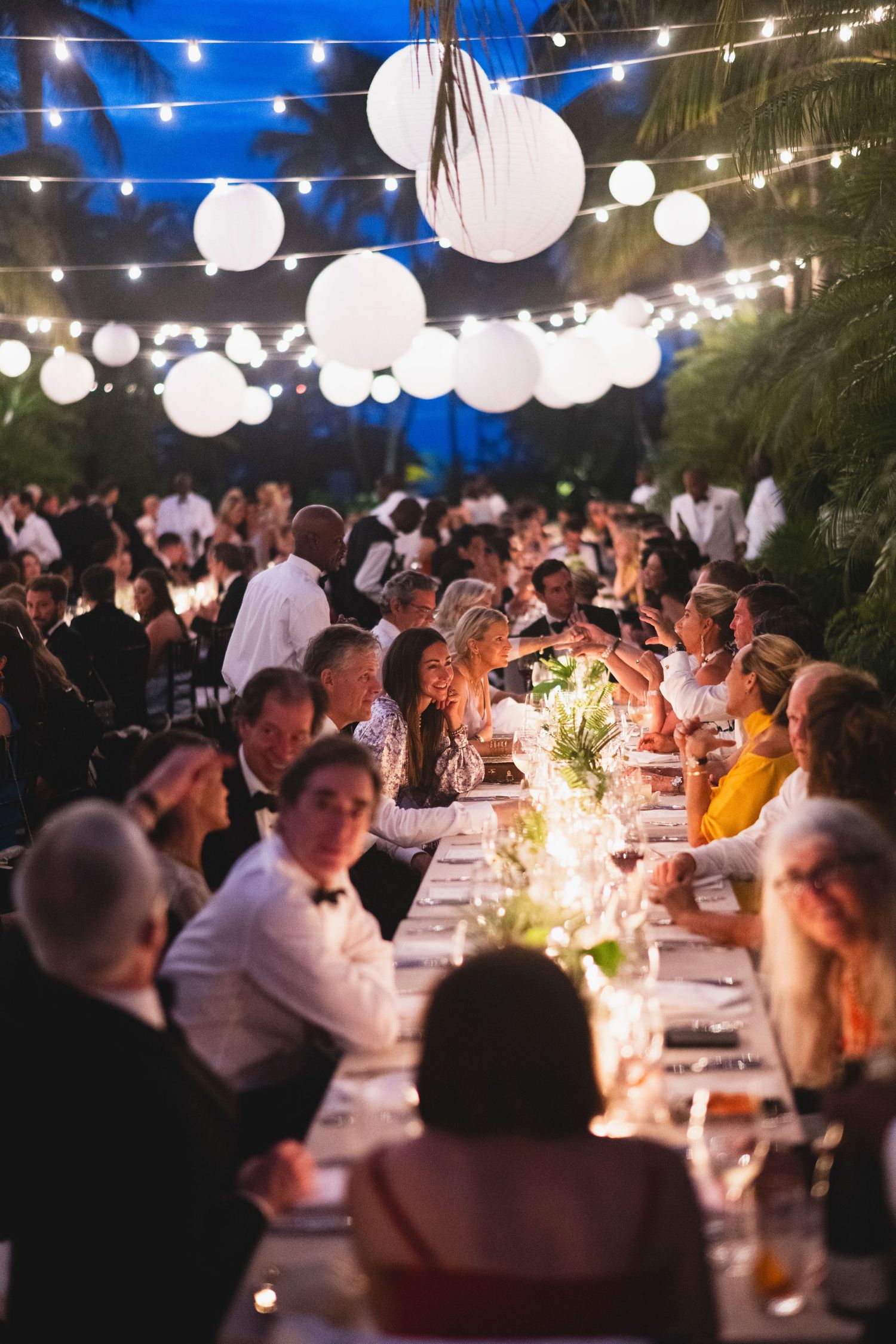 Guests enjoy an elegant outdoor evening wedding reception under glowing string lights and paper lanterns at dusk.