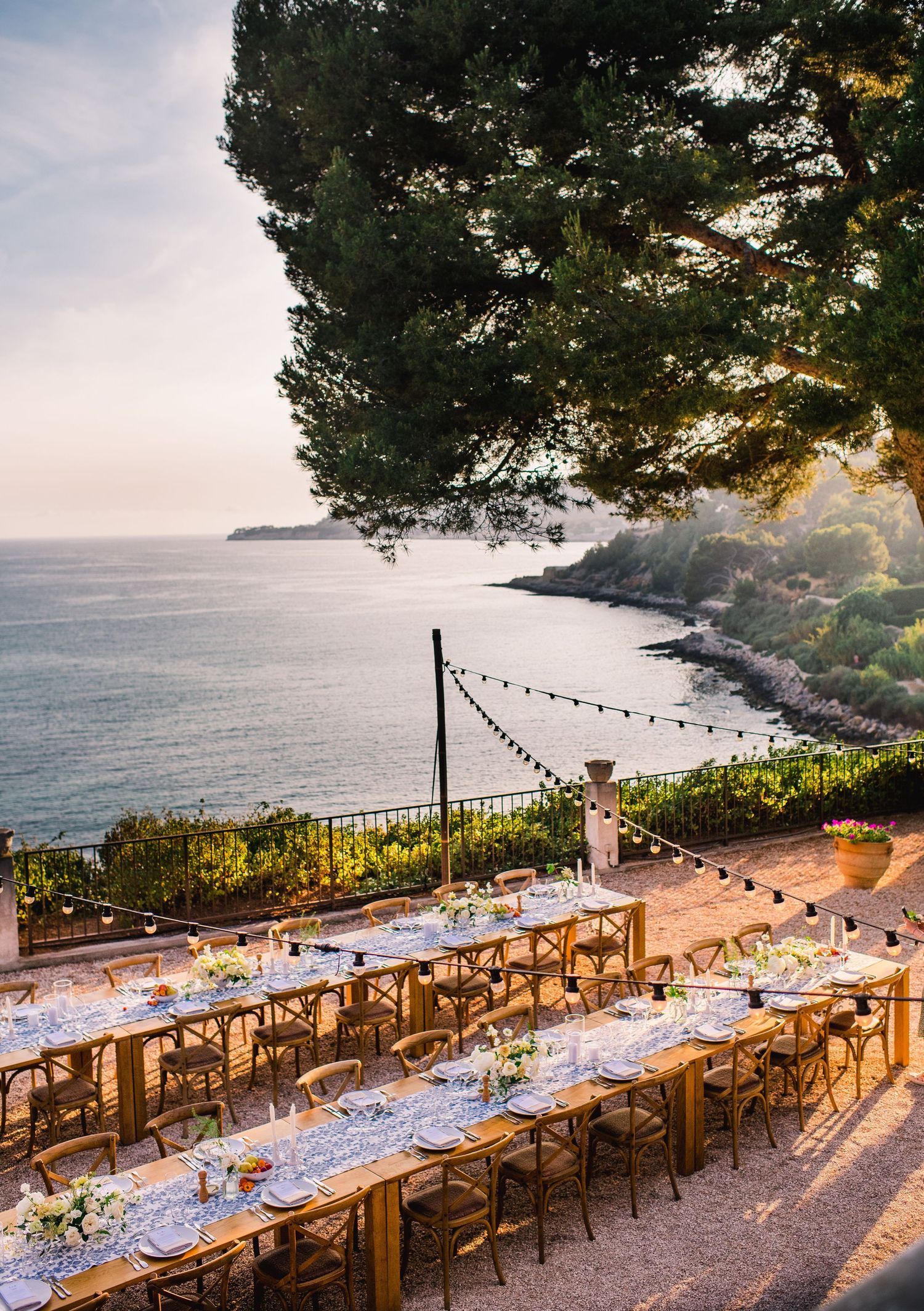 Long rustic wooden dining tables set up for an outdoor wedding reception overlooking a scenic Mediterranean coastal view at sunset.