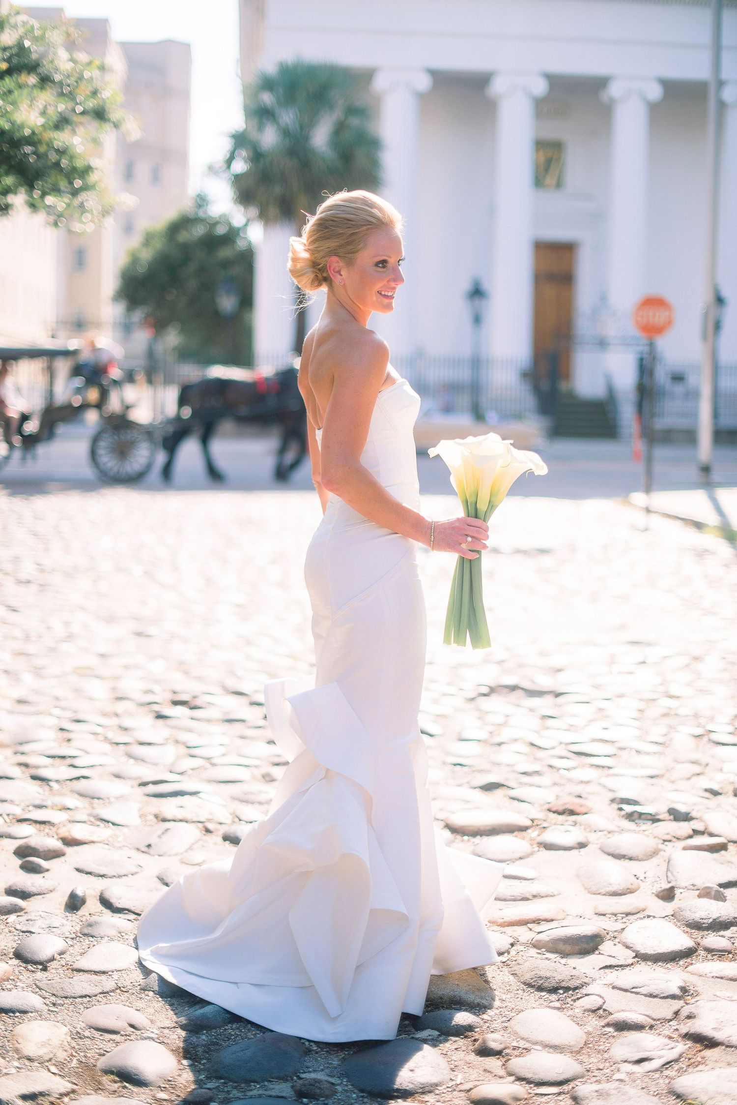 A bride in a white wedding dress sits gracefully on cobblestone steps holding a yellow bouquet in outdoor photos.