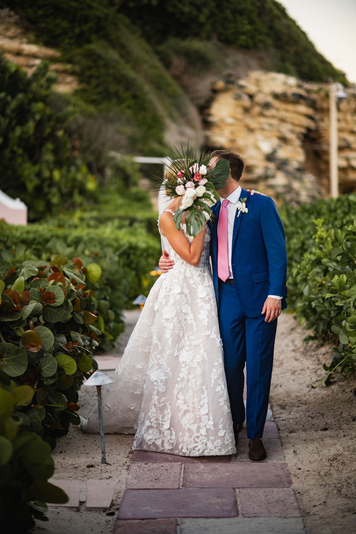 Couple in wedding attire walking along stone pathway surrounded by tropical foliage.