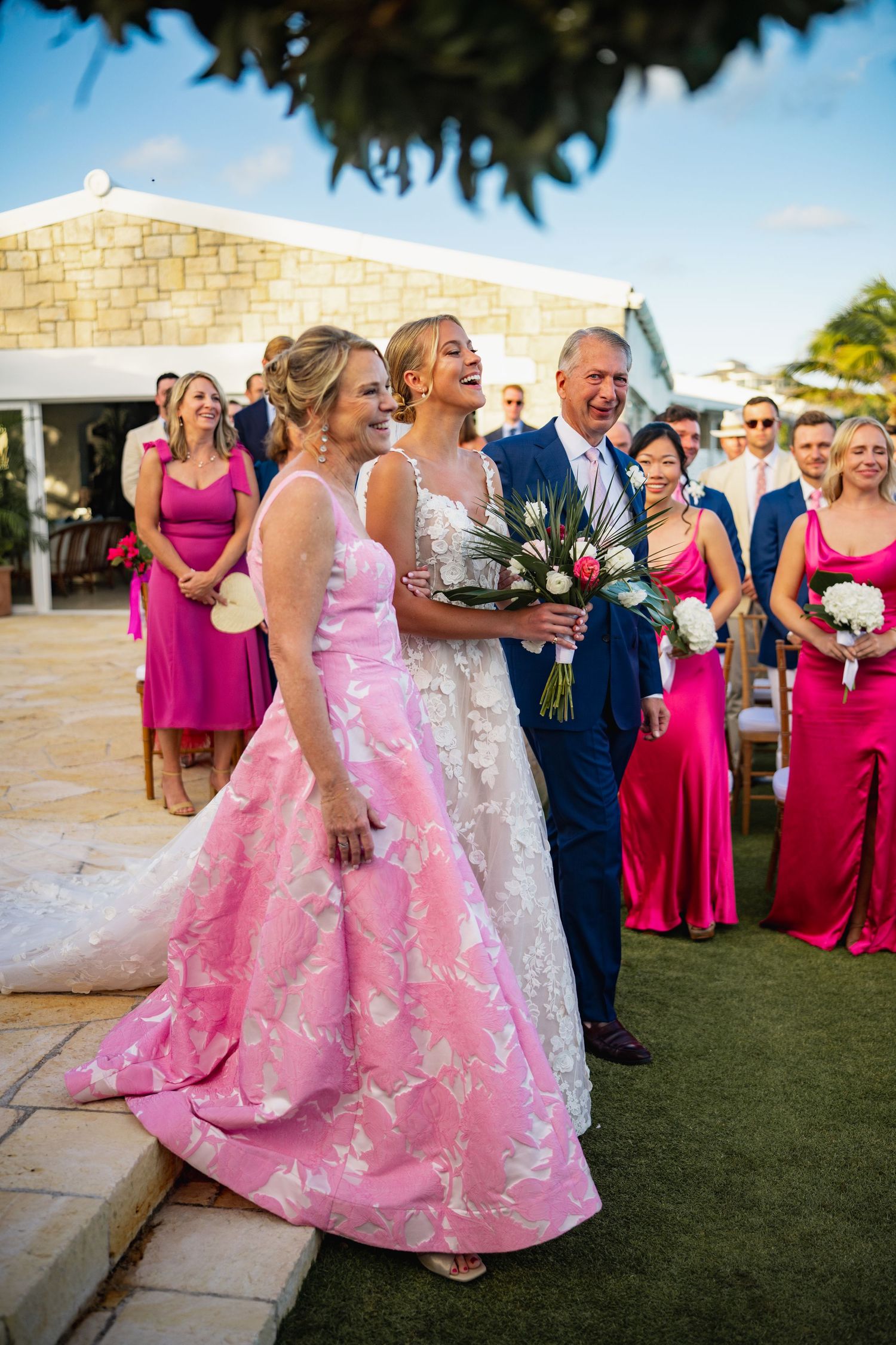 Wedding party in pink and magenta dresses gathers on a stone patio under blue skies.