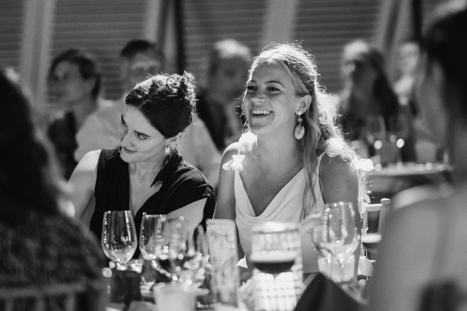 Wedding guests share joyful laughter during an elegant reception dinner with wine glasses on the table.