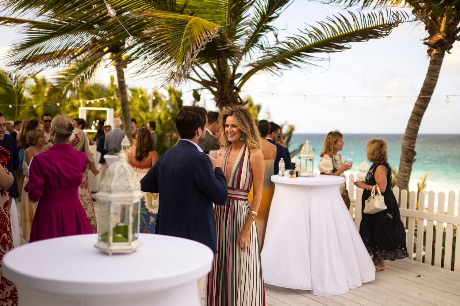 Beachside wedding reception in Harbour Island with palm trees and ocean views as guests mingle around cocktail tables.