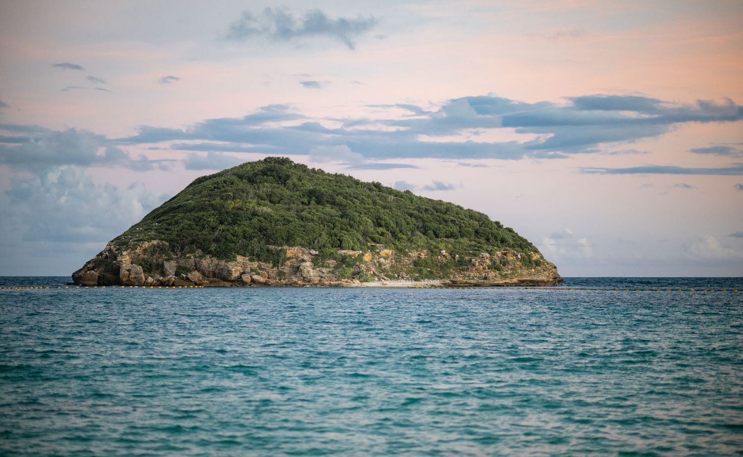 Small dome-shaped island rising from calm blue waters of Antigua at the Mill Reef Club during sunset in the Caribbean.
