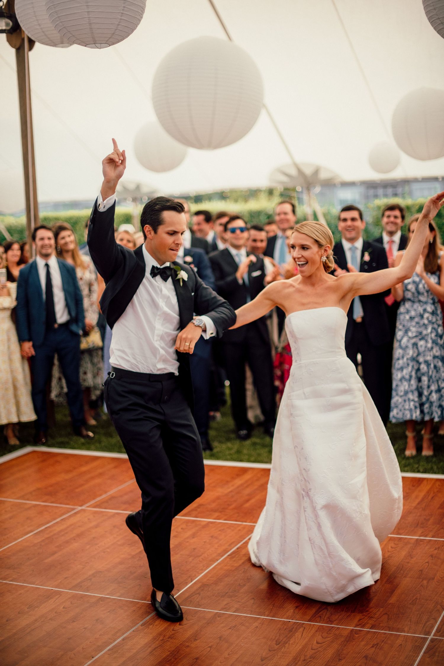 Joyful dancing celebration on wooden dance floor under white paper lanterns at wedding reception.