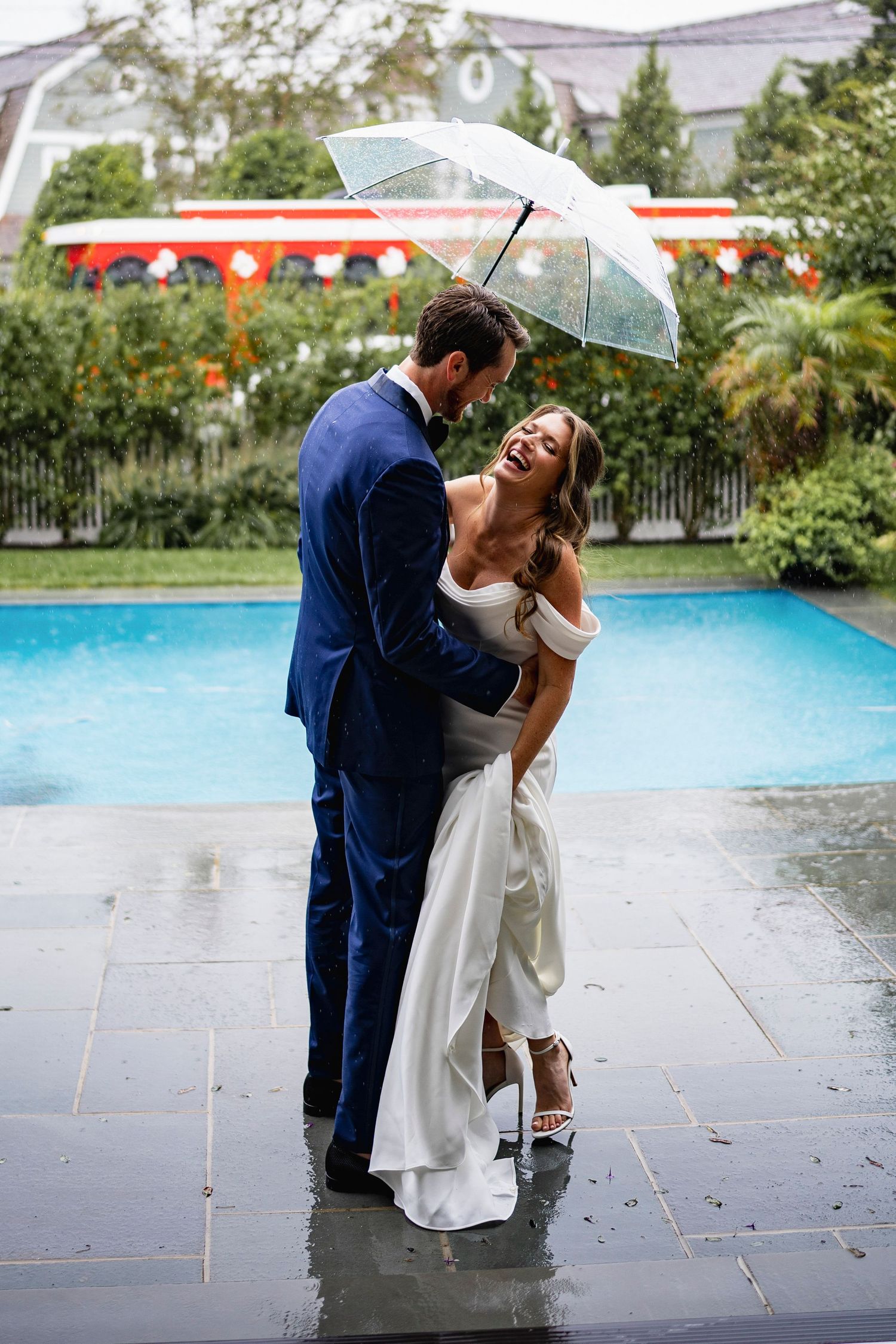 Couple shares romantic moment under clear umbrellas on rainy wedding day by pool.