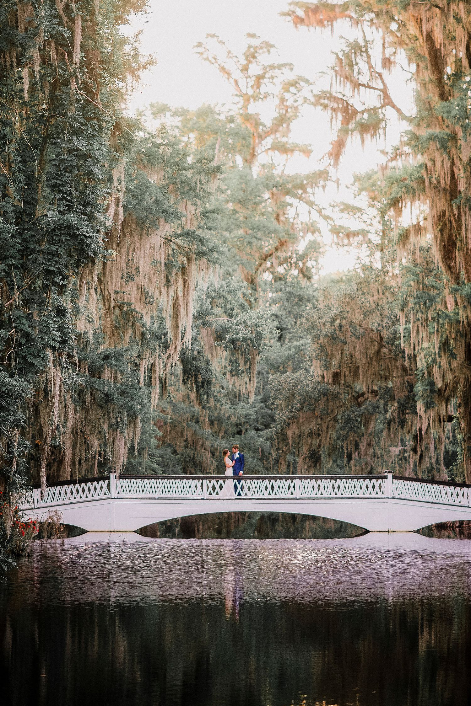 A white ornate bridge spans across a tranquil pond surrounded by Spanish moss-draped trees in a southern garden setting.