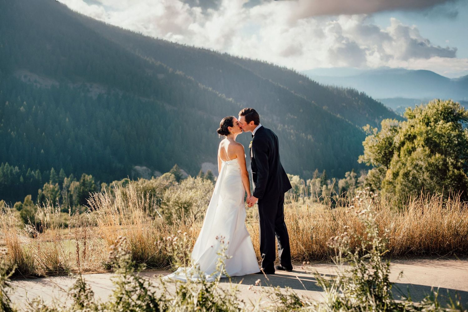 A couple shares a romantic moment on a mountain overlook with sunlit hills and dramatic clouds in the background.