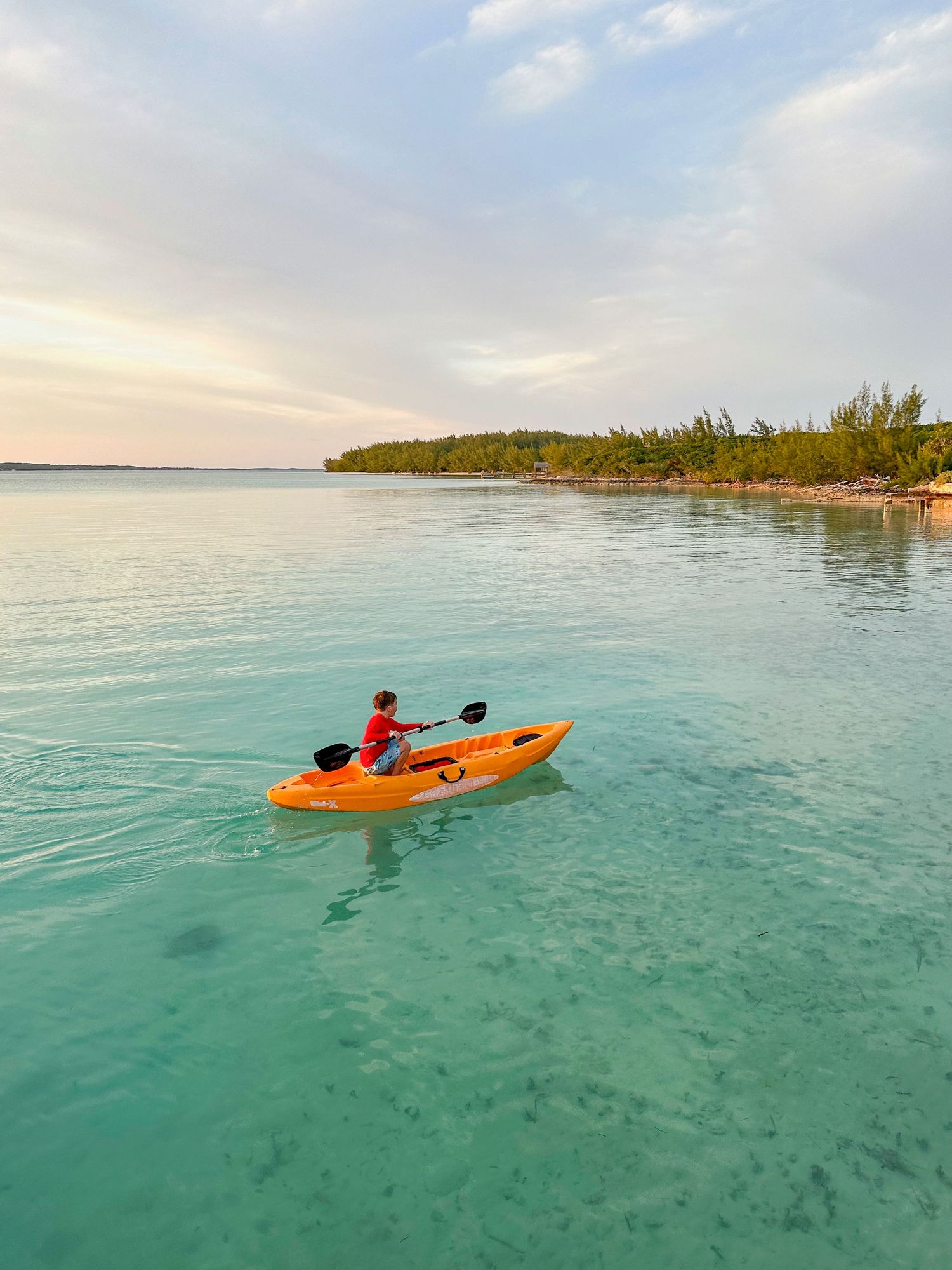 Orange kayak glides through crystal clear turquoise waters near a tropical shoreline during sunset.