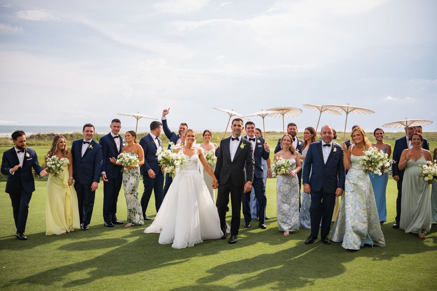 Wedding party members stand in a line on a grassy hill with blue sky and clouds in the background.