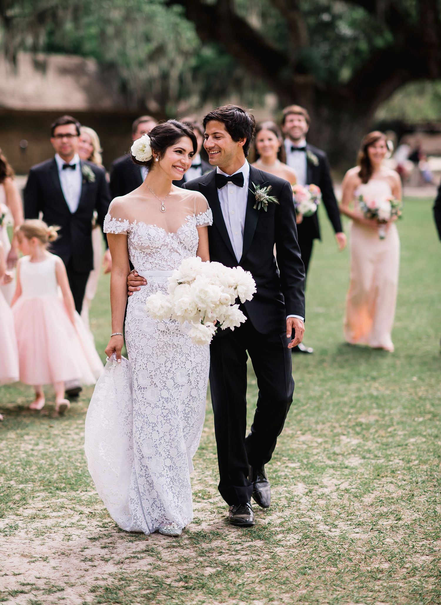 Wedding party walks together on grassy lawn in formal attire with bridesmaids in pink dresses and groomsmen in black suits.