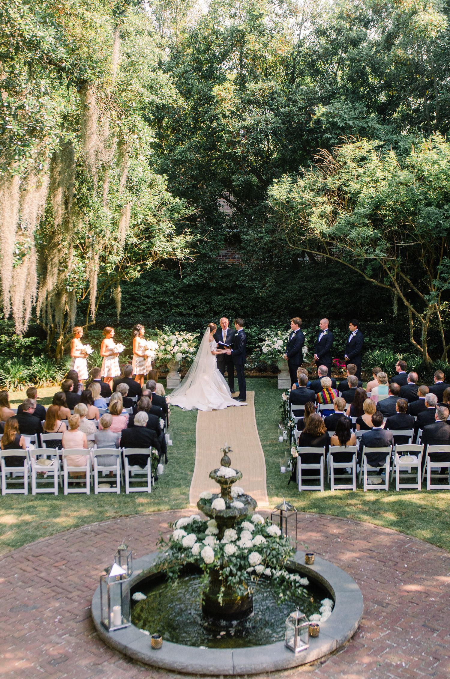 Outdoor garden wedding ceremony setup with white chairs arranged in rows on a brick patio surrounded by lush greenery.