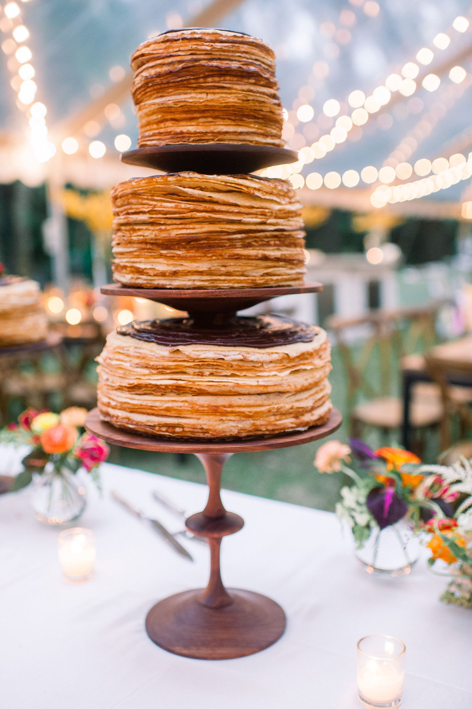 A stack of crepe cakes on brown cake stands decorated with string lights and flowers in the background.