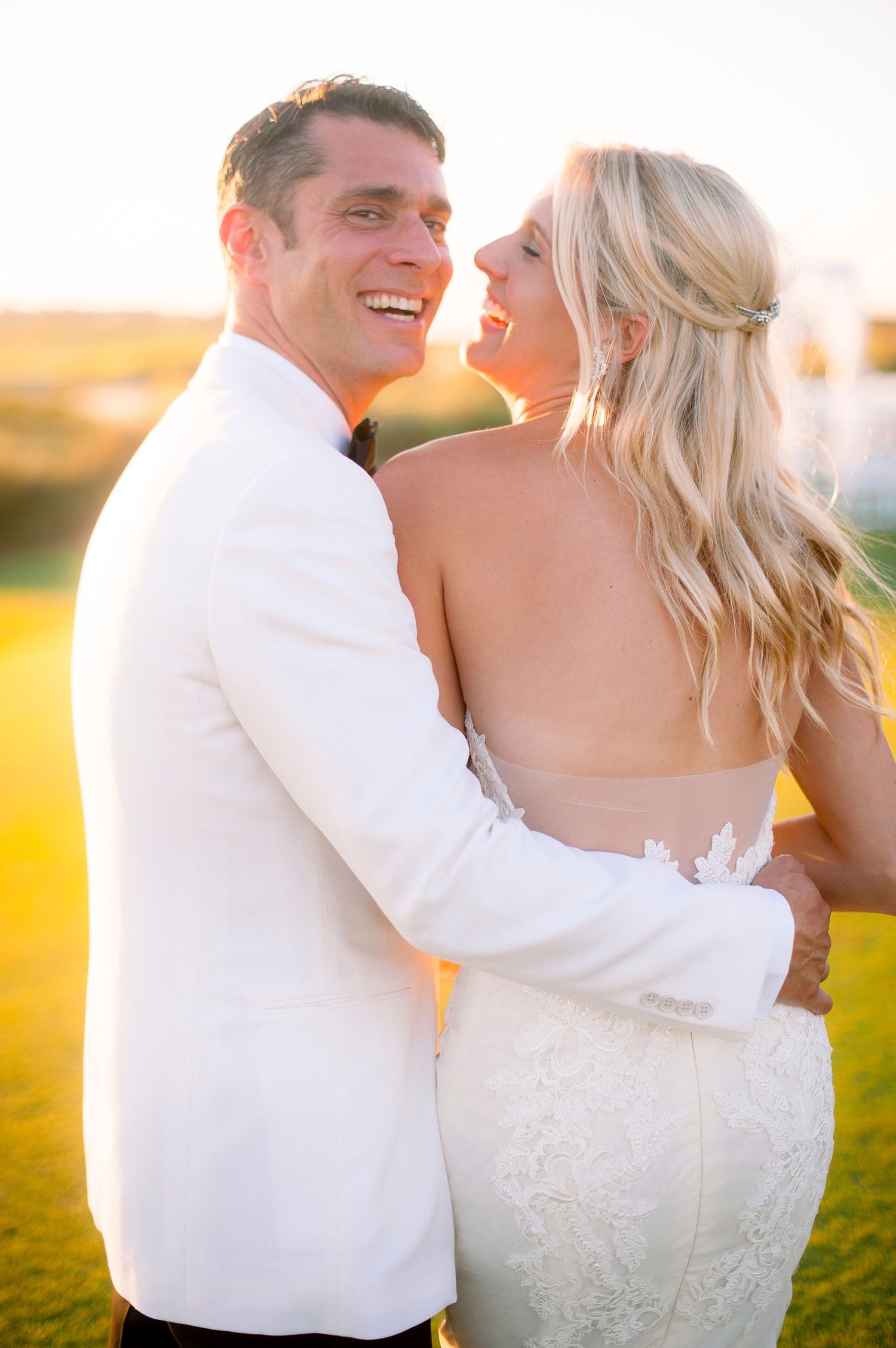 A couple in wedding attire embraces and laughs together in a sunlit field during golden hour.