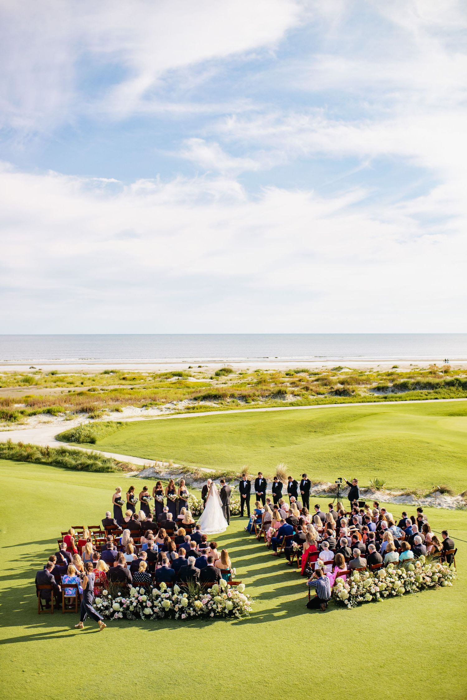 Aerial view of wedding guests gathered on a lush green golf course with ocean views and blue skies in the background.